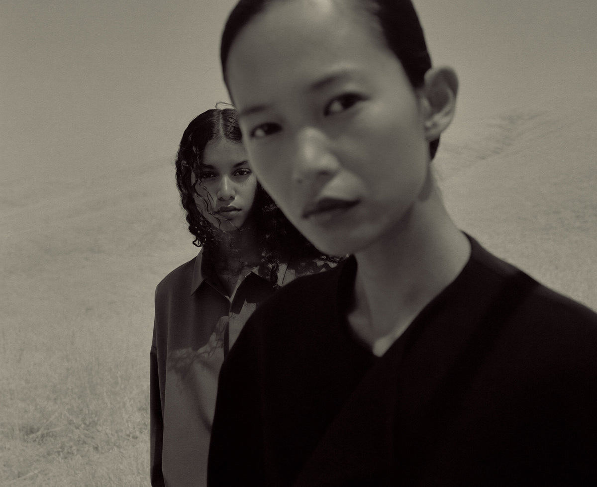 Monochrome portrait of a person in a dark V neck top standing close to the camera, with a second person in layered clothing visible against a textured neutral backdrop