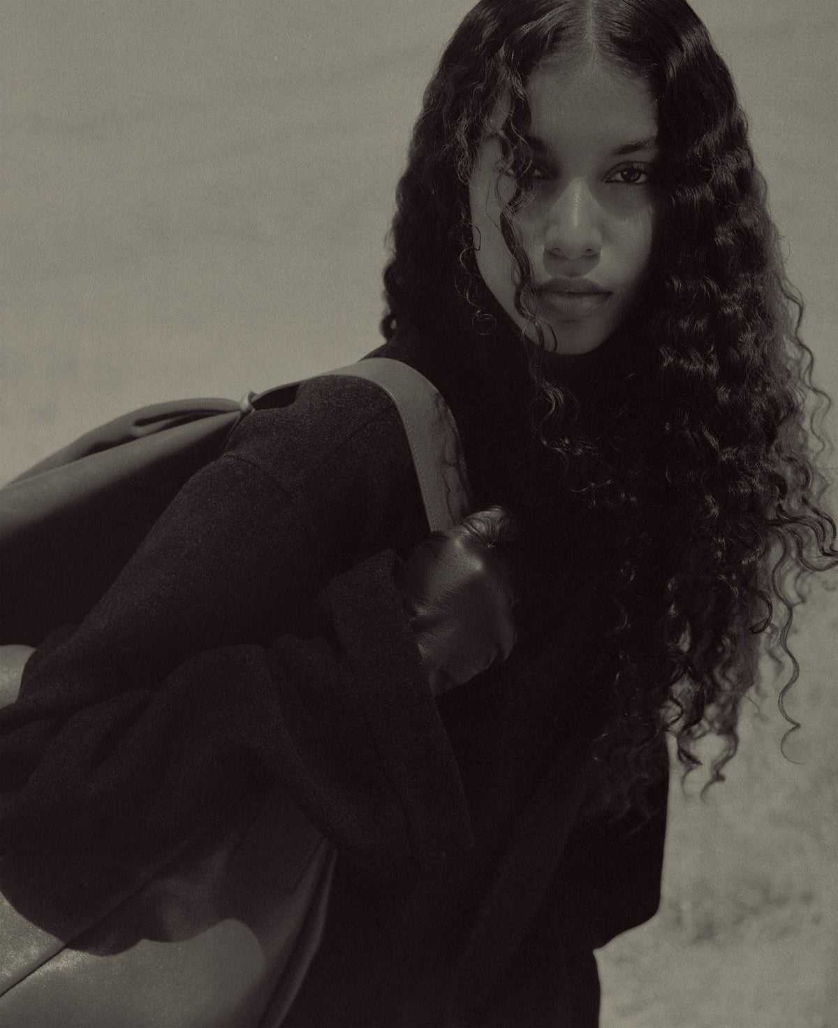 Monochrome portrait of a woman with long curly hair wearing dark Outerwear and carrying a shoulder bag against a softly textured backdrop