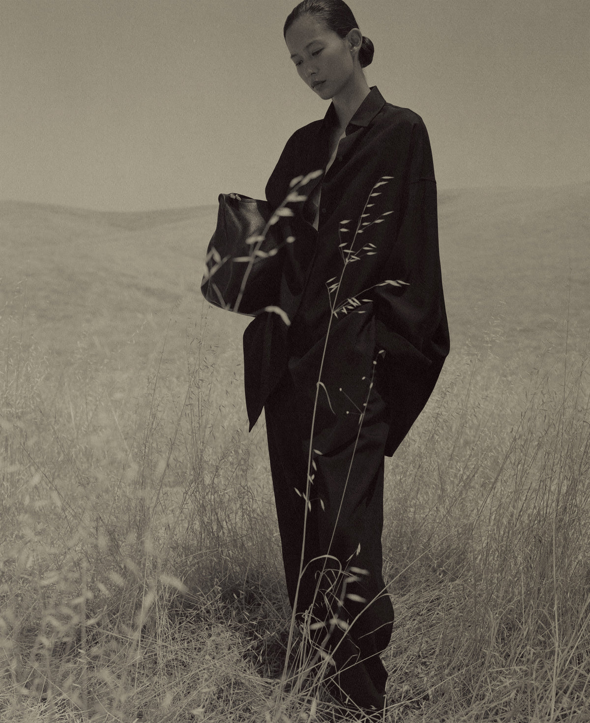 Sepia portrait of a woman in dark suiting standing among tall dry grass, holding a black leather bag against a softly rolling landscape