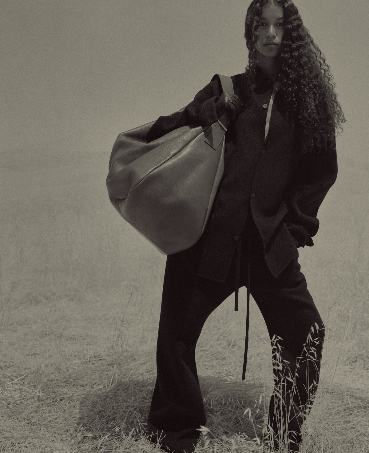 Monochrome portrait of a woman with long curly hair wearing dark suiting and carrying an oversized shoulder bag in a grassy field