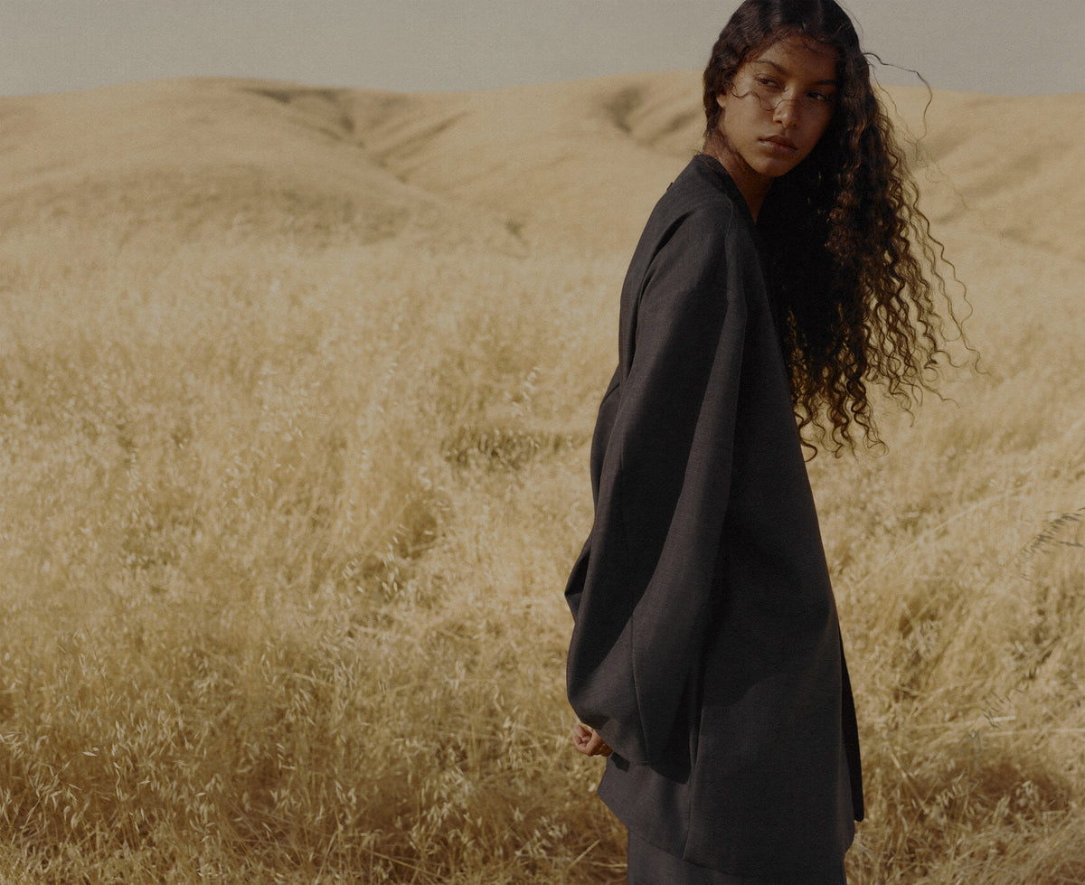 Woman with long curly hair wearing dark oversized Outerwear while standing in a sunlit golden grass field with softly rolling hills behind her