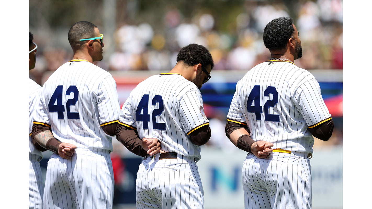 Three baseball players in white pinstriped uniforms with the number 42 on their backs stand side by side with hands behind their backs during a game.
