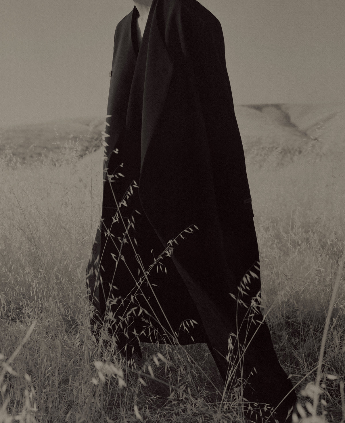 Monochrome figure wrapped in a long black cloak, standing among tall grasses and wildflowers in a muted open field under an overcast sky