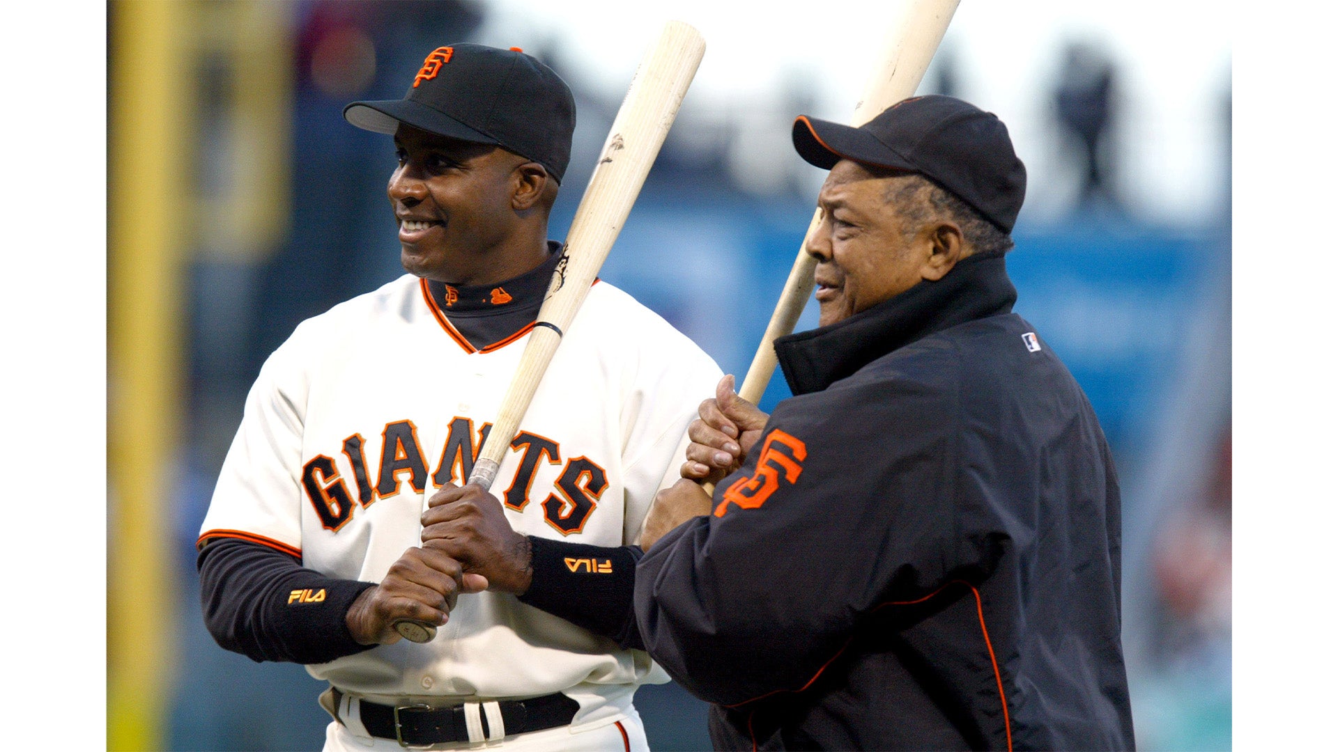 Two men wearing San Francisco Giants baseball gear stand side by side on a field, smiling and holding wooden baseball bats during daylight.