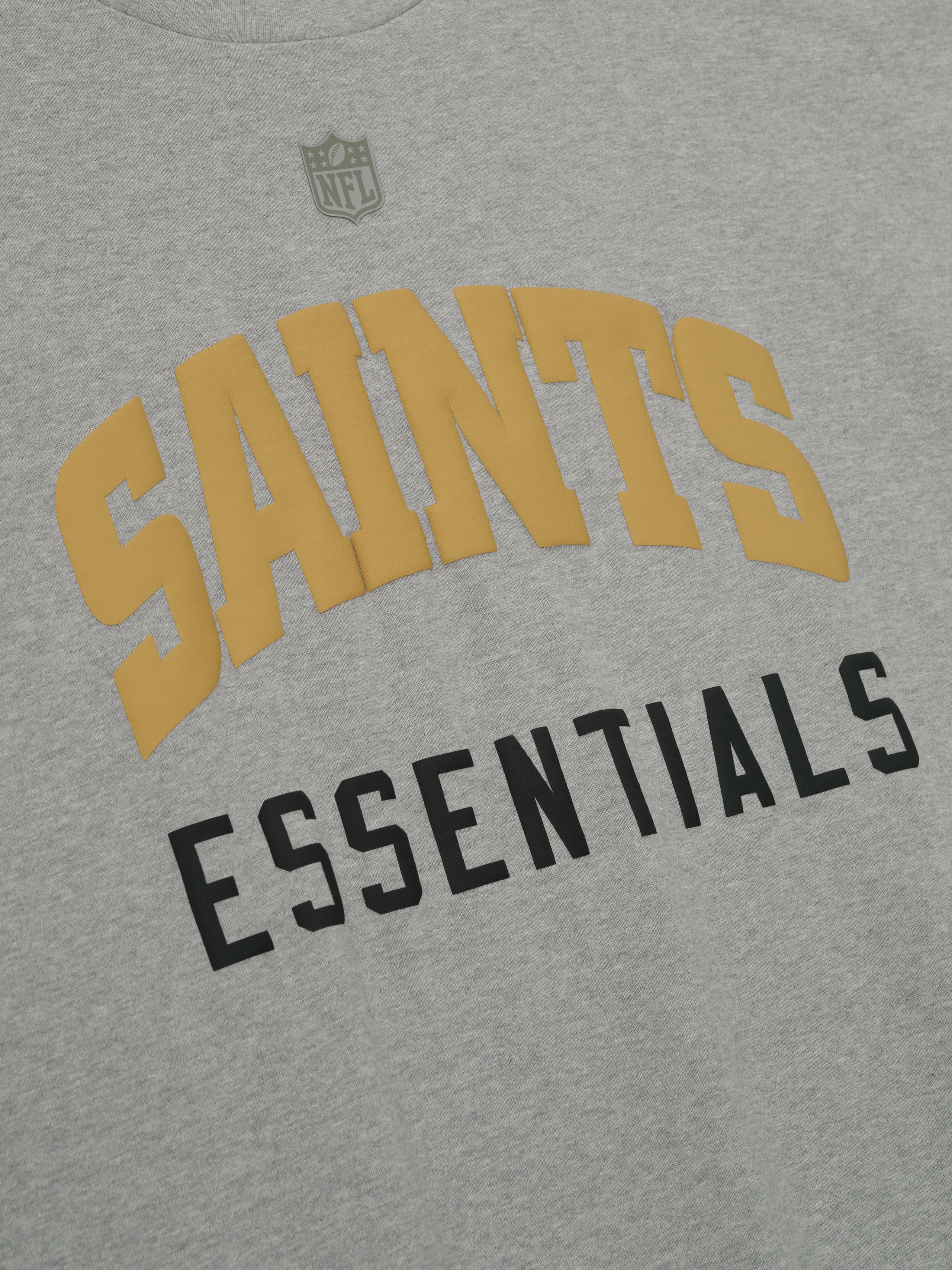 Close-up of concrete heather Saints 90s 3/4 Sleeve Football Tee showing gold Saints arched text and black Essentials below center chest
