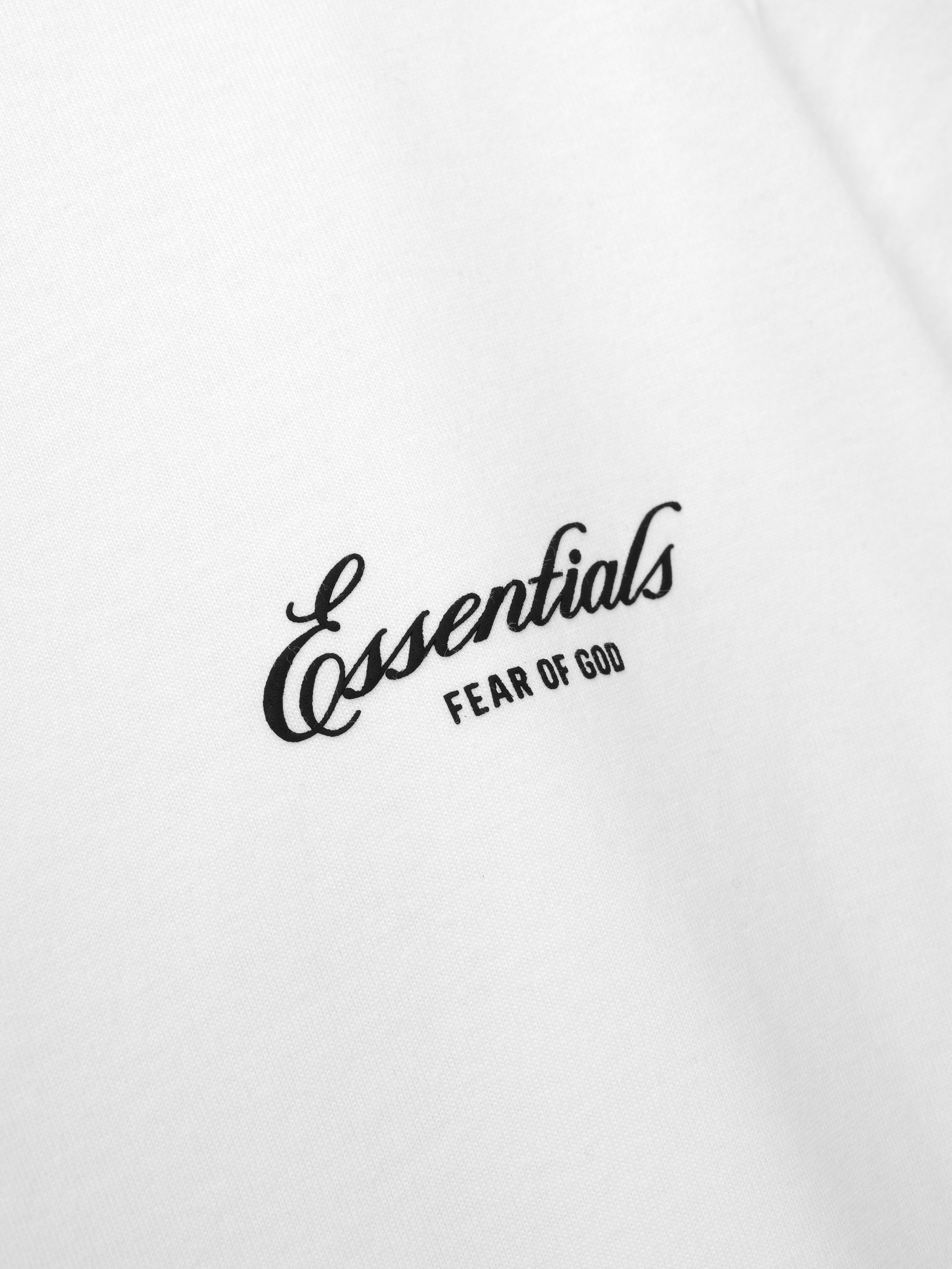 Close-up of the white Signature SS26 Classic Tee displaying black cursive "Essentials" and small "Fear of God" text branding on soft cotton fabric.