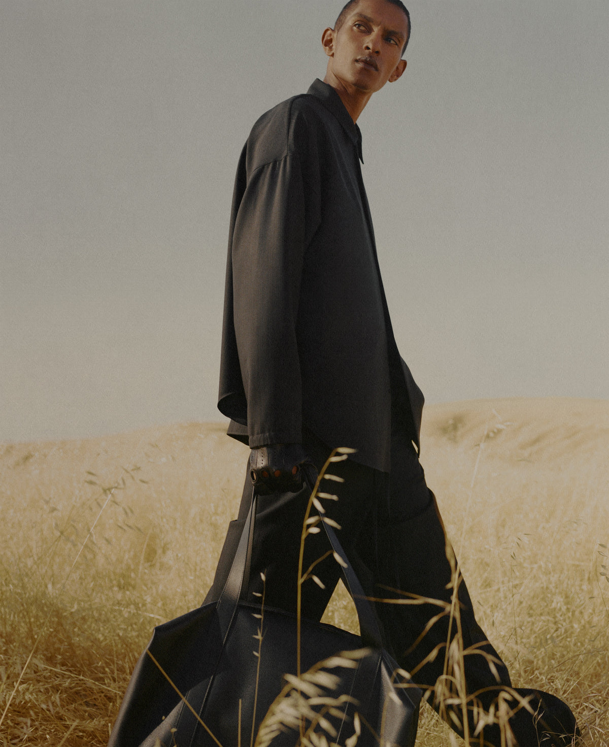 Man in flowing black Outerwear standing among tall golden grass, with a muted gray sky and rolling dry field behind him in a softly lit portrait