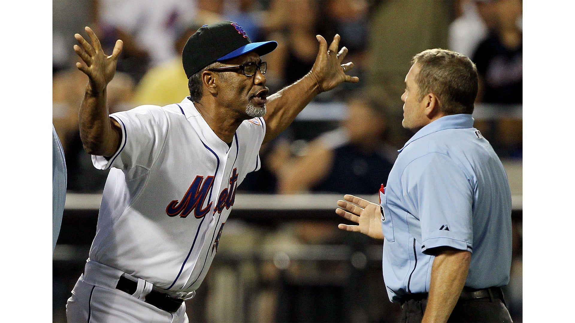 A baseball coach in a white Mets uniform with glasses and a cap gestures passionately with arms wide open while speaking to an umpire wearing a light blue shirt during a game.