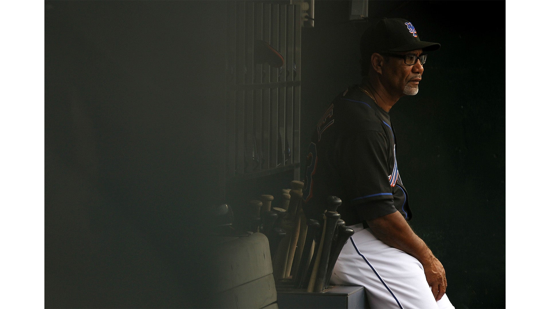 An older man wearing a black baseball jersey and cap sits quietly on a bench in a dimly lit dugout with several bats leaning against the wall beside him.