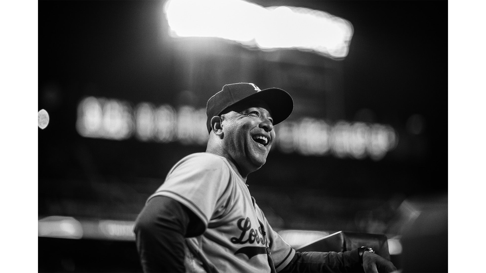 Black and white image of a smiling baseball player wearing a cap and jersey standing in a stadium with blurred lights in the background.