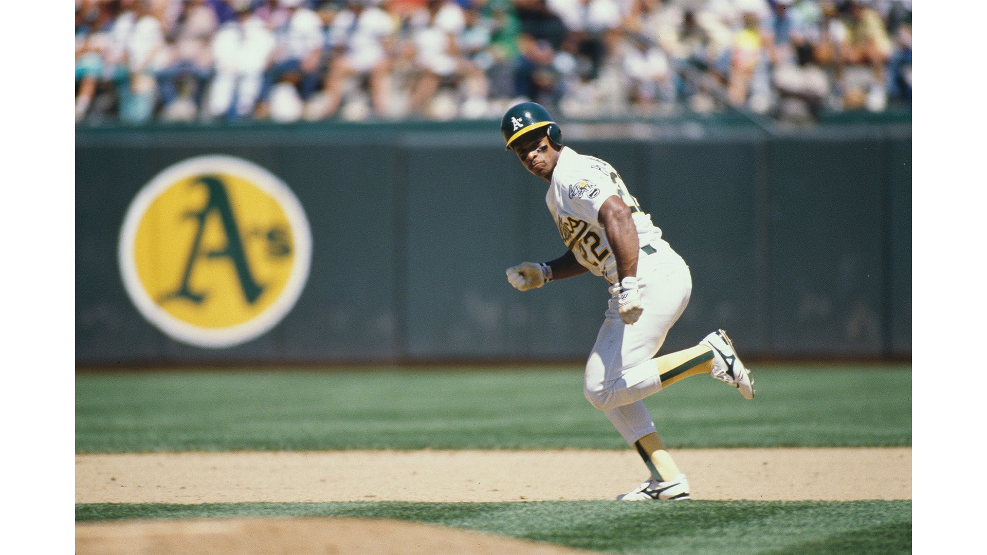 A baseball player wearing a white Oakland Athletics uniform with number 12 runs on the field during a daytime game, with the team's logo visible on the outfield wall behind him.