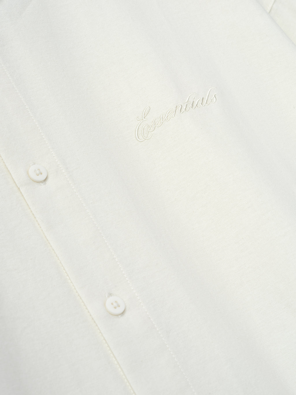 Close-up of ivory fabric with tonal embroidered "Essentials" logo and small buttons along the front placket