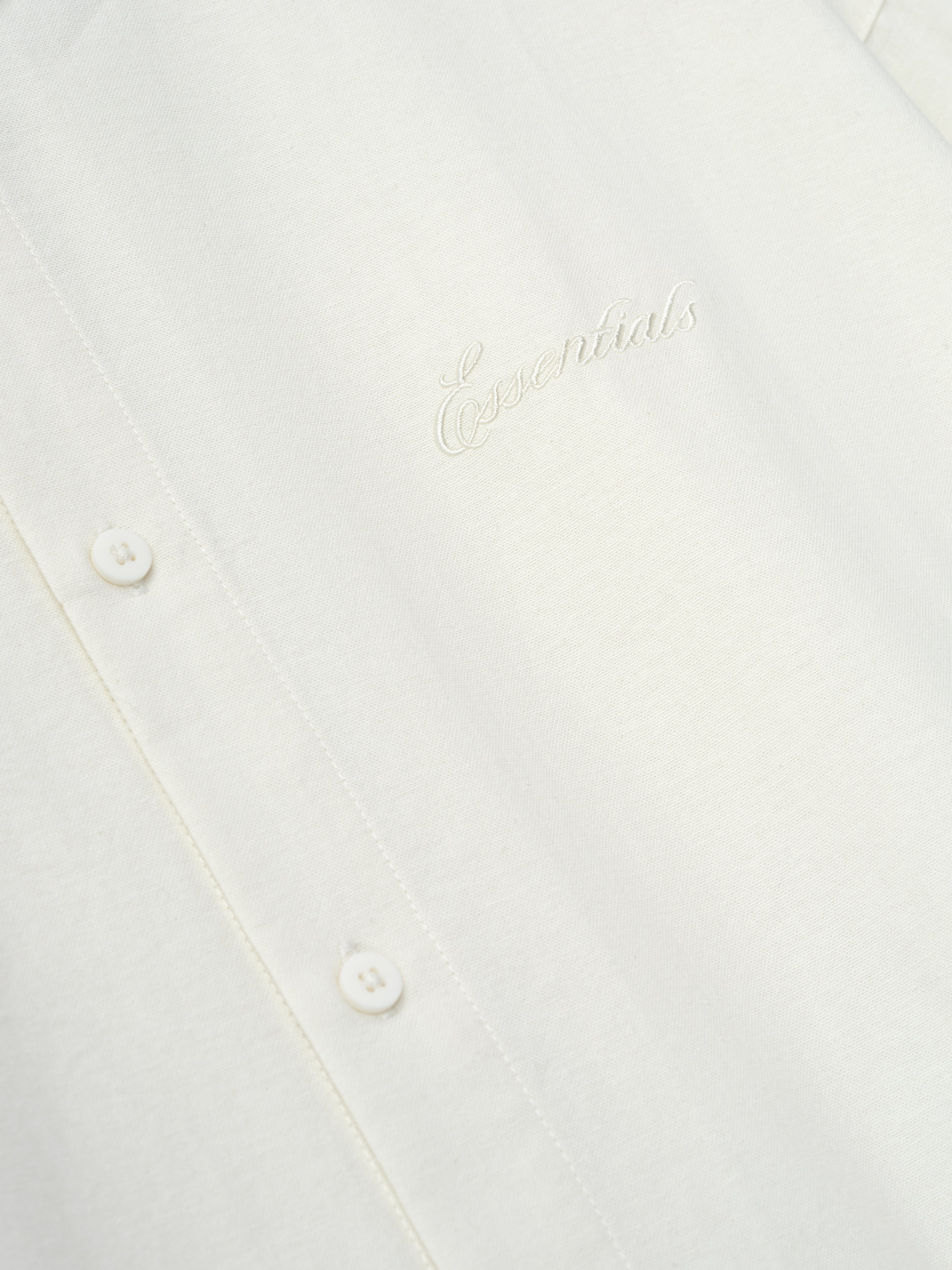 Close-up of ivory fabric with tonal embroidered "Essentials" logo and small buttons along the front placket