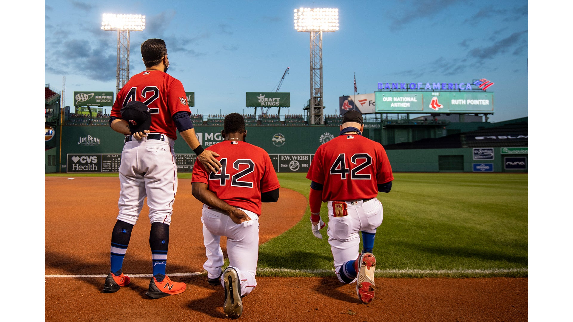 Three baseball players wearing red jerseys with the number 42 on the back kneel and stand together on the field near the infield dirt at a stadium during early evening.