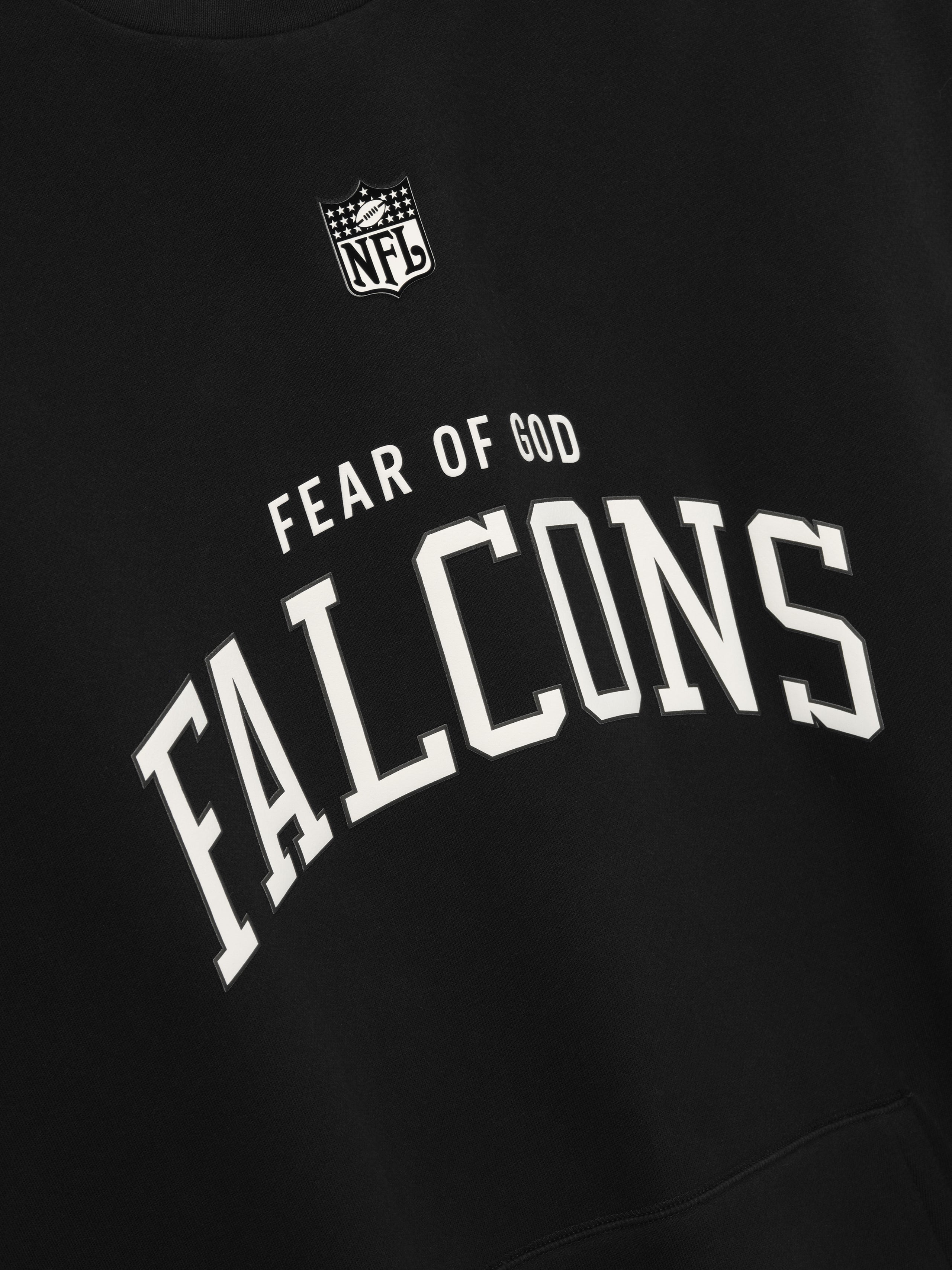 Black crewneck with white arched "FALCONS" text and small "FEAR OF GOD" above NFL logo on front fabric close-up