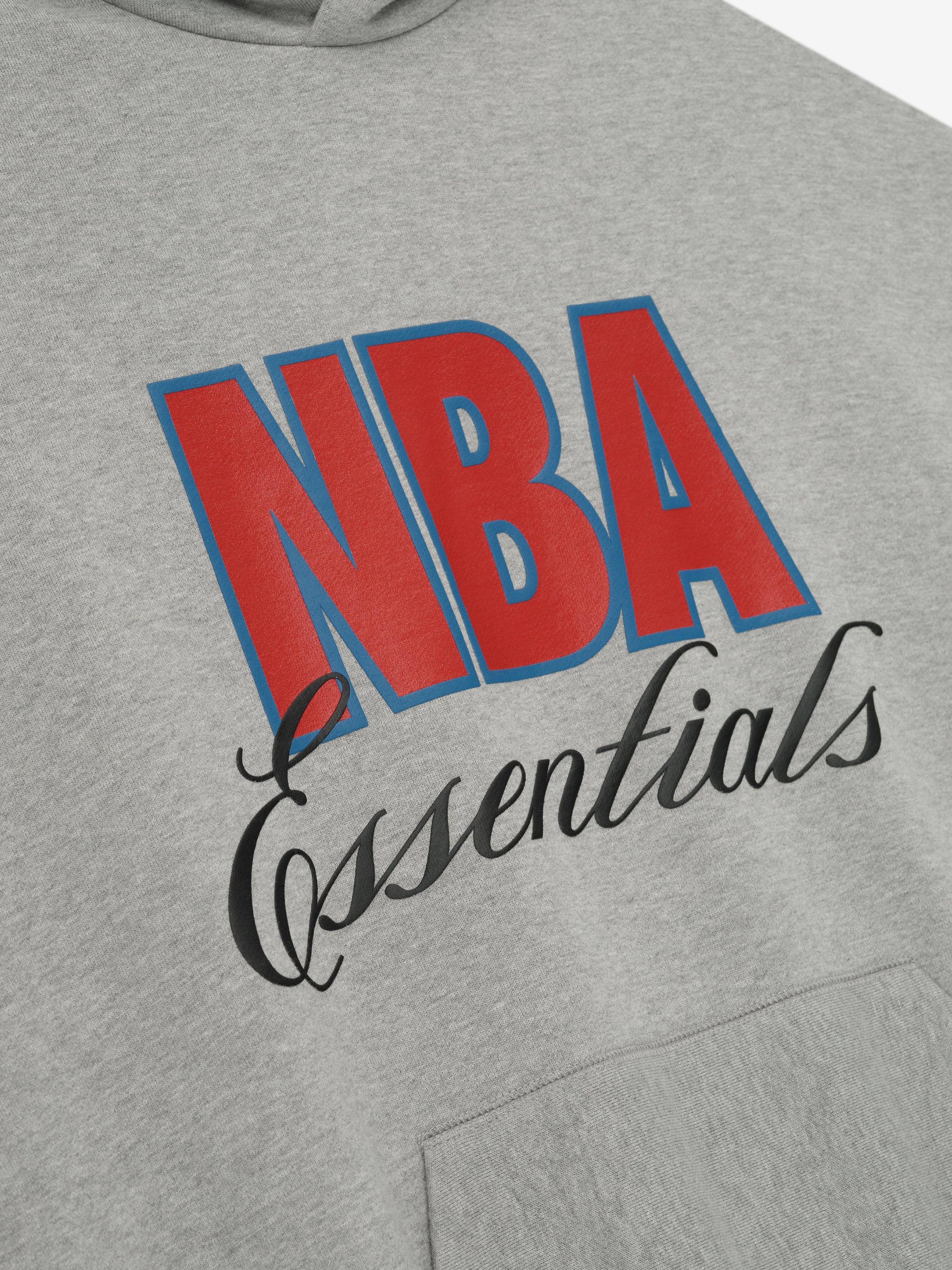 Close-up of concrete heather NBA 90s hoodie front showing bold red and blue NBA text with black "Essentials" script above kangaroo pocket