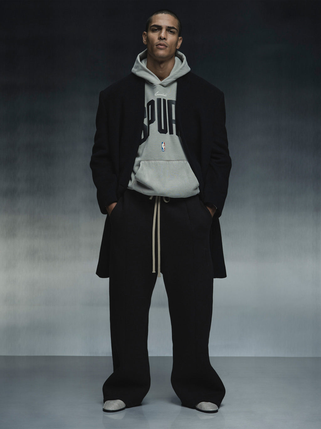 Model wearing grey Spurs 90s Hoodie layered under black coat and wide-leg pants, full front view against dark backdrop