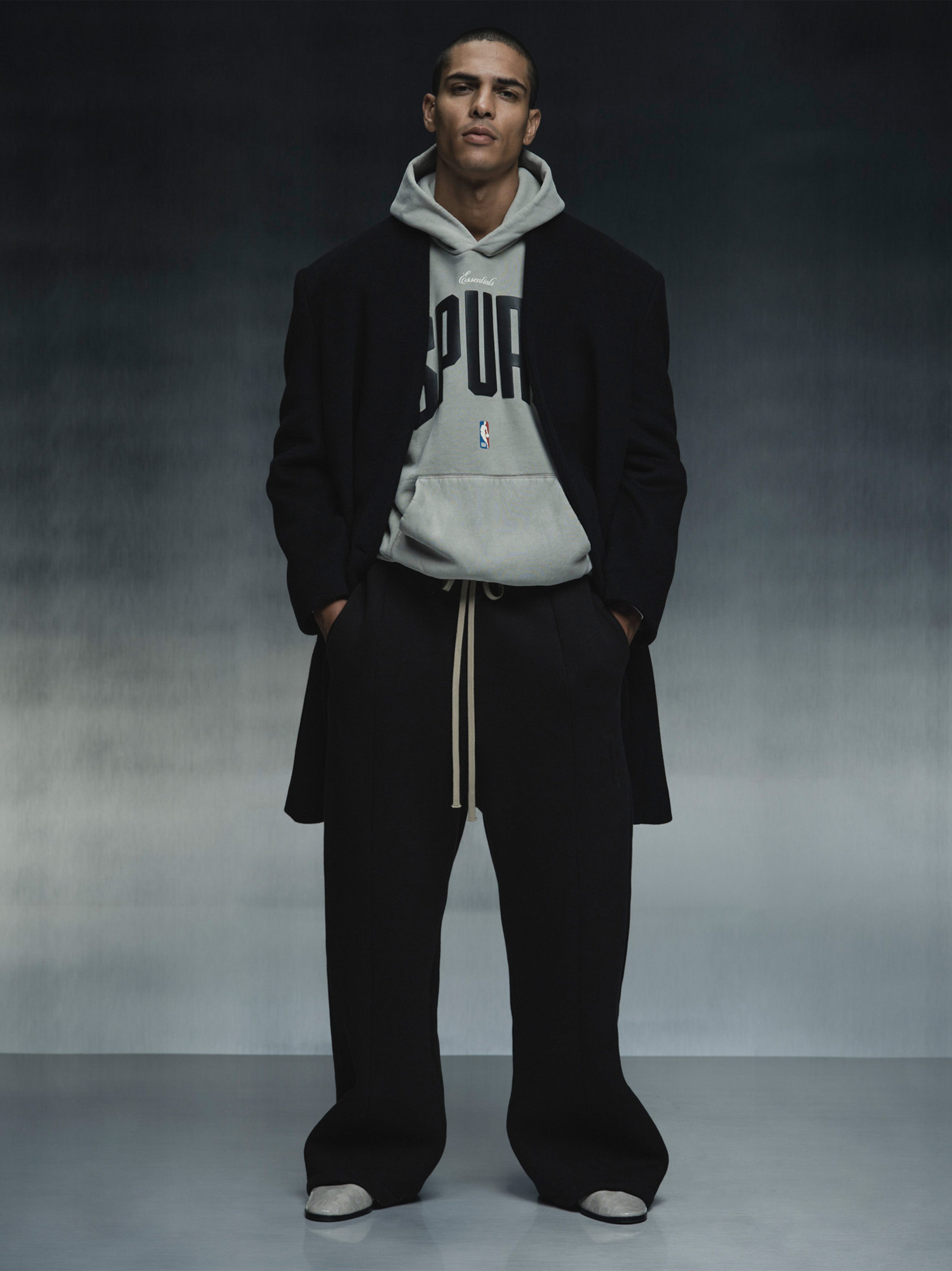 Model wearing grey Spurs 90s Hoodie layered under black coat and wide-leg pants, full front view against dark backdrop