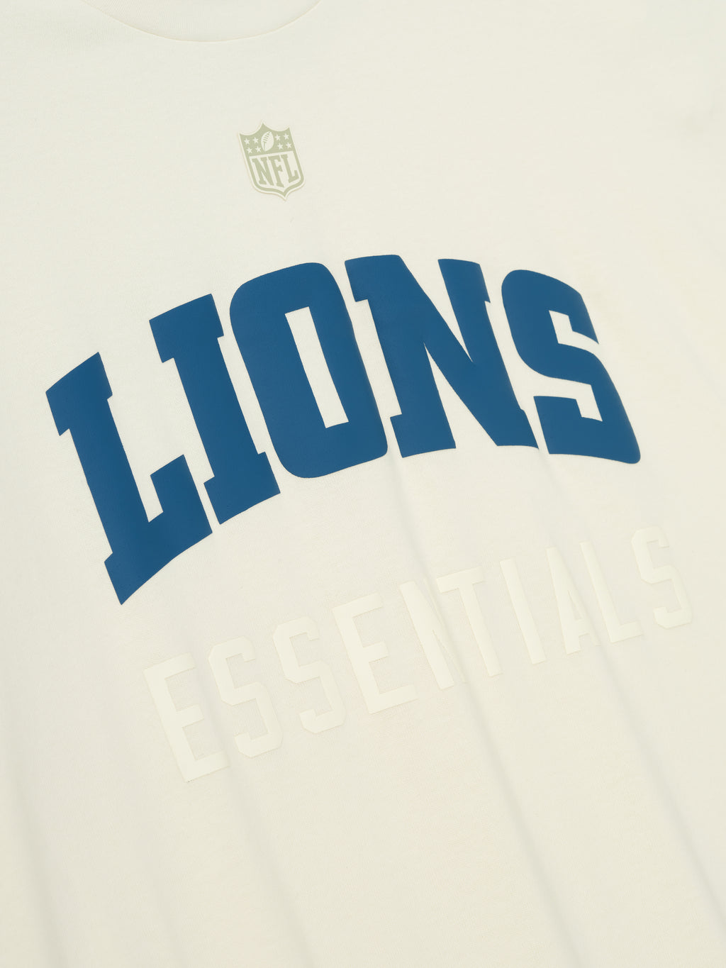 Close-up of cream white Lions 90s 3/4 Sleeve Football Tee showing bold blue Lions text and subtle Essentials lettering below