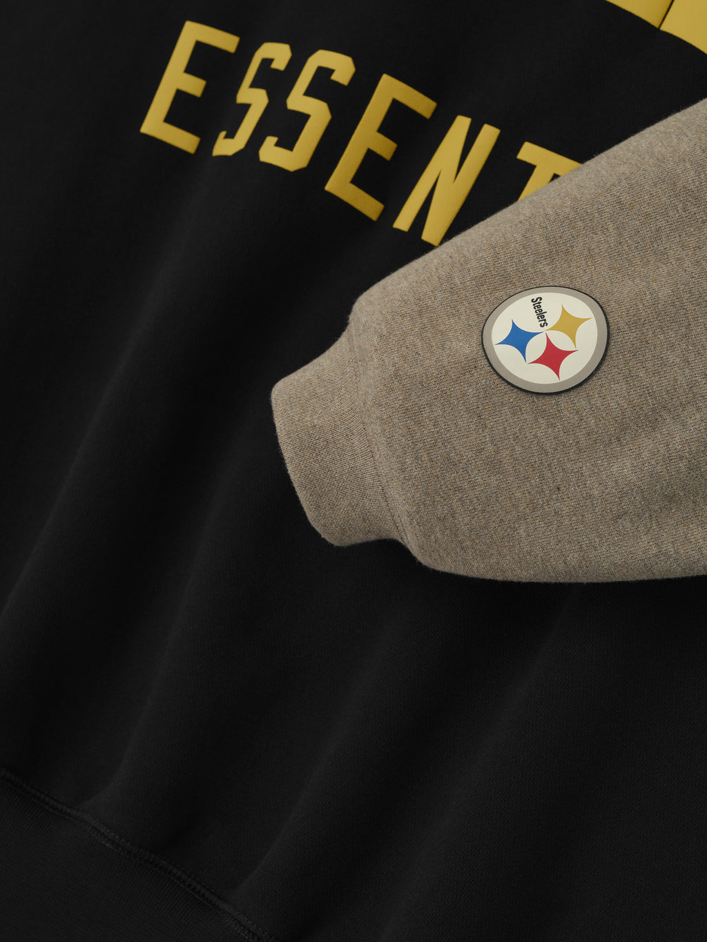 Close-up of Steelers Sport Hoodie showing black body with gold Essentials text and heather grey sleeve with Steelers logo patch, gallery 0