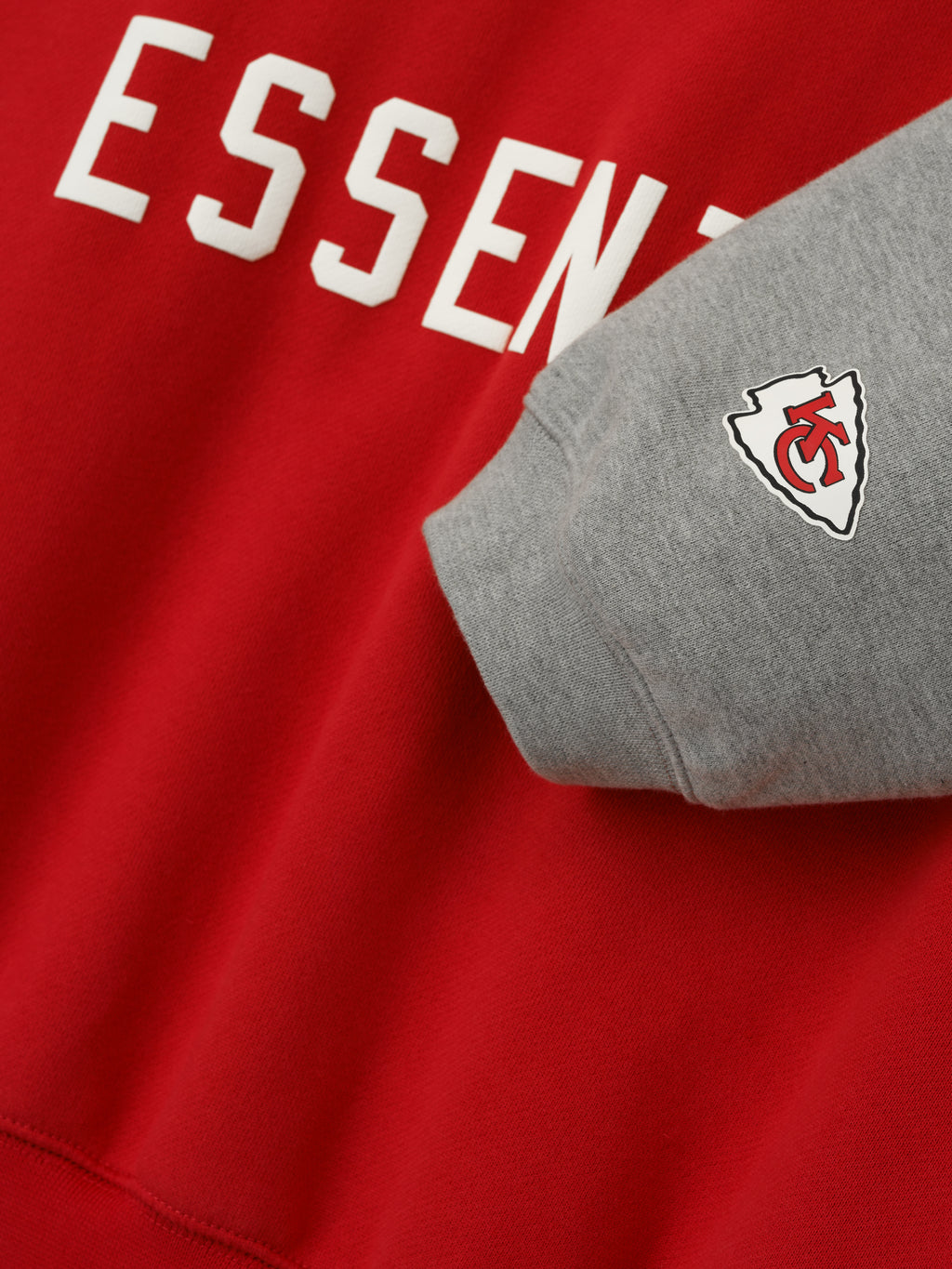 Close-up of Chiefs Sport Hoodie showing red body with white Essentials text and contrasting concrete heather sleeve with team logo patch