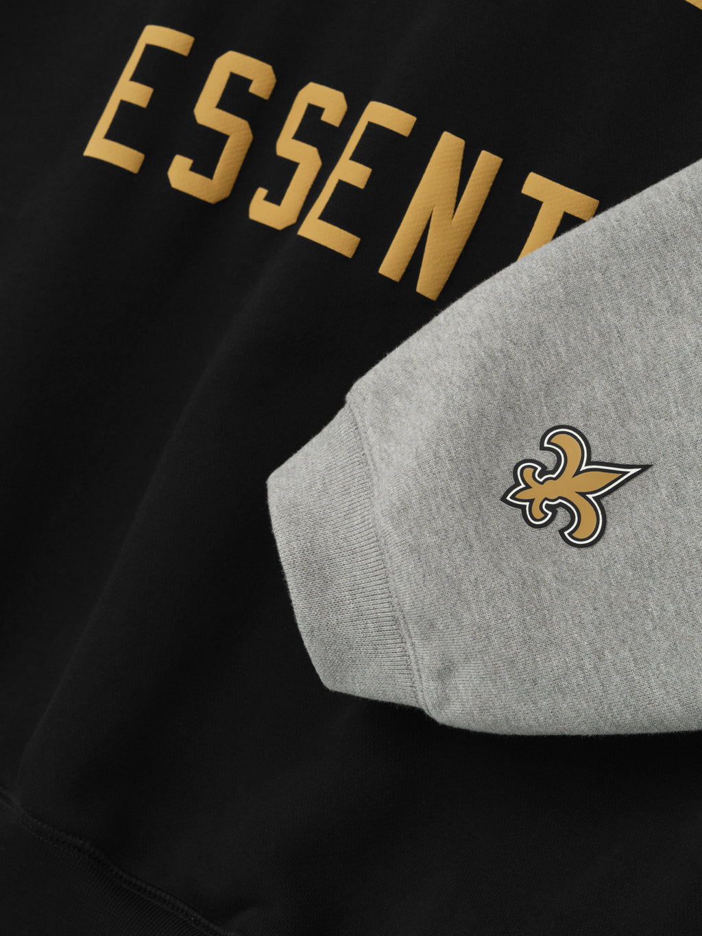 Close-up of Saints Sport Hoodie showing partial gold Essentials text on black body and grey sleeve with fleur-de-lis logo patch