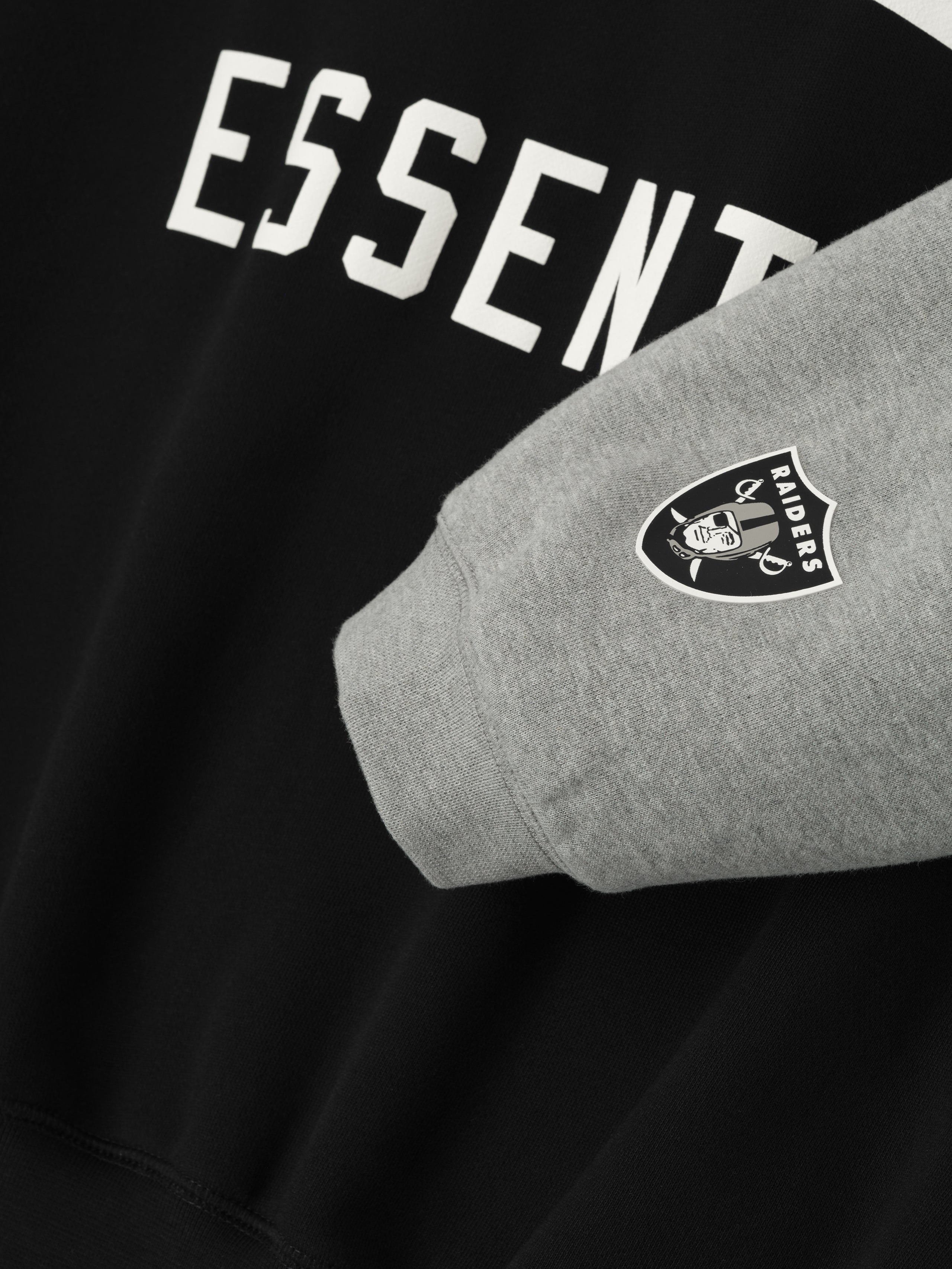 Close-up of Raiders Sport Hoodie showing concrete heather sleeve with team logo and black body with partial Essentials text