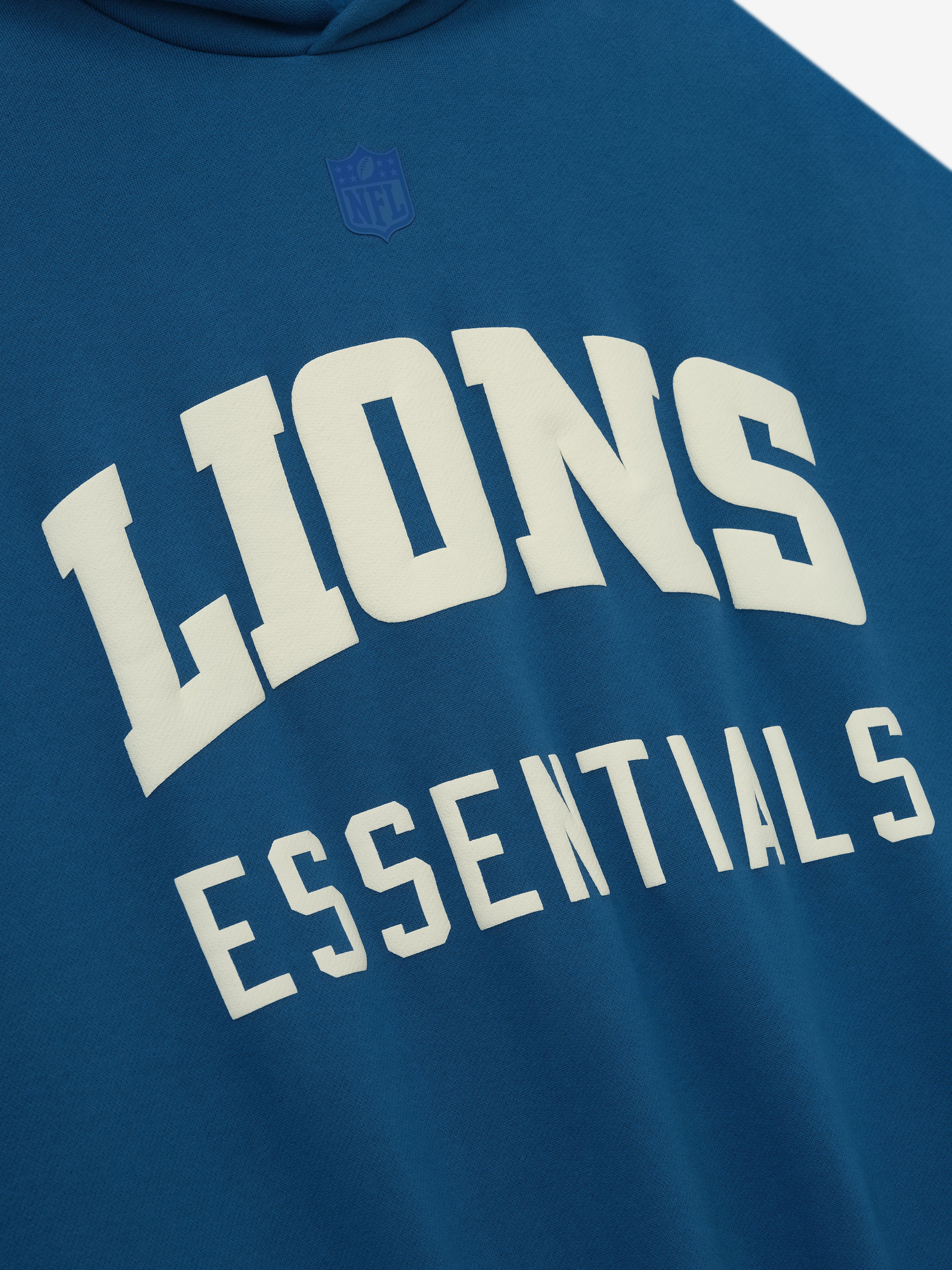 Close-up of blue sapphire Lions Sport Hoodie front showing large white Lions Essentials text and subtle tone-on-tone NFL logo above