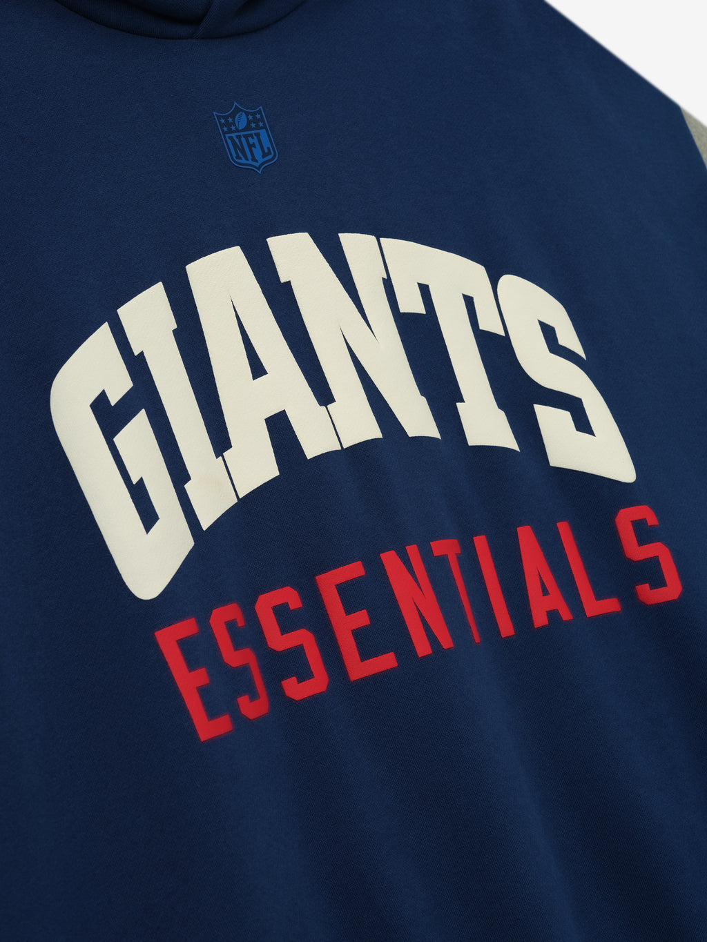 Close-up of Giants Sport Hoodie in blue estate fabric showing white Giants text and red Essentials lettering beneath NFL shield logo
