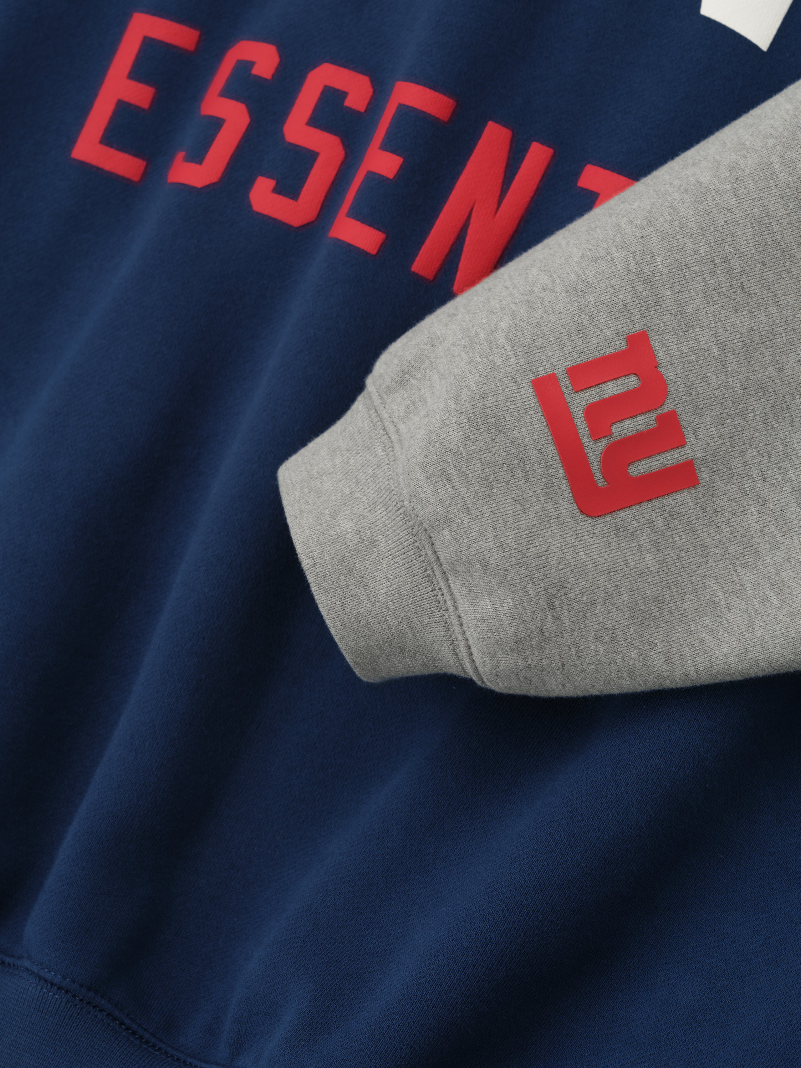 Close-up on concrete heather sleeve with red NY Giants logo and navy body featuring red Essentials text on hoodie