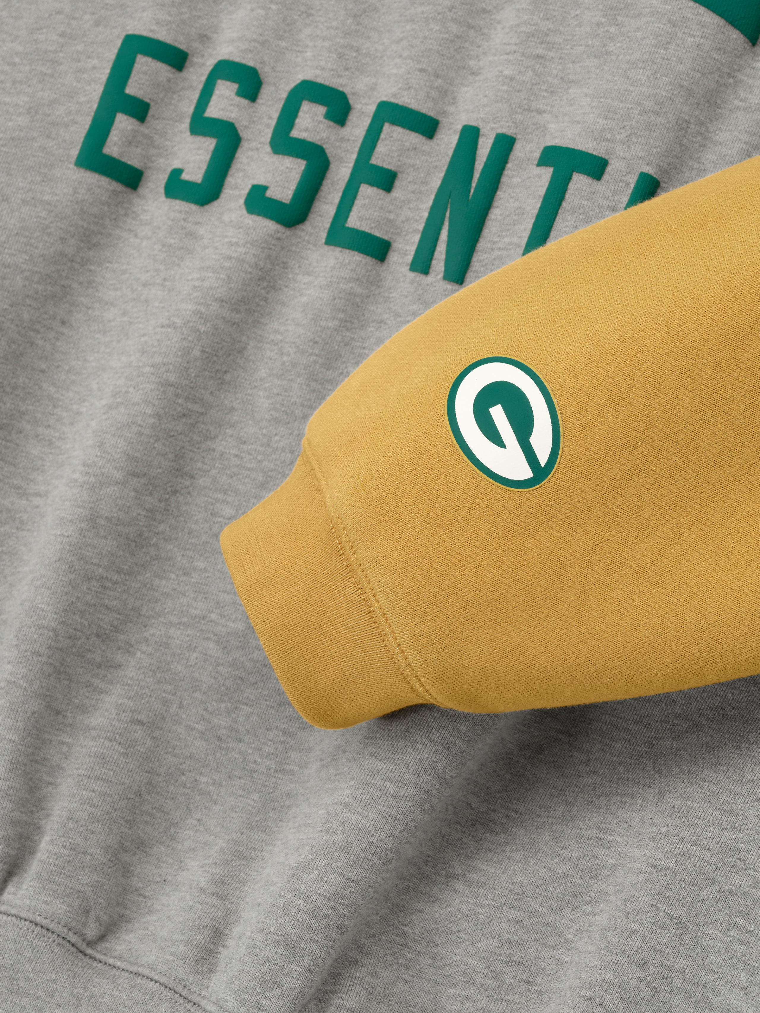 Close-up of Packers Sport Hoodie in concrete heather showing green Essentials text and canyon sleeve with team logo patch