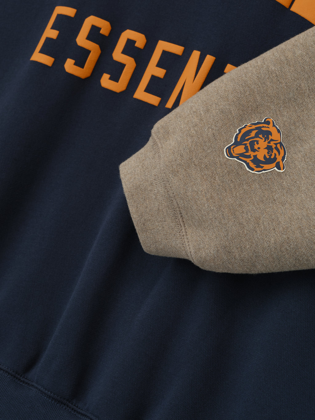 Close-up of Bears Sport Hoodie showing orange Essentials text on dark sapphire fabric and gray sleeve with small bear logo patch