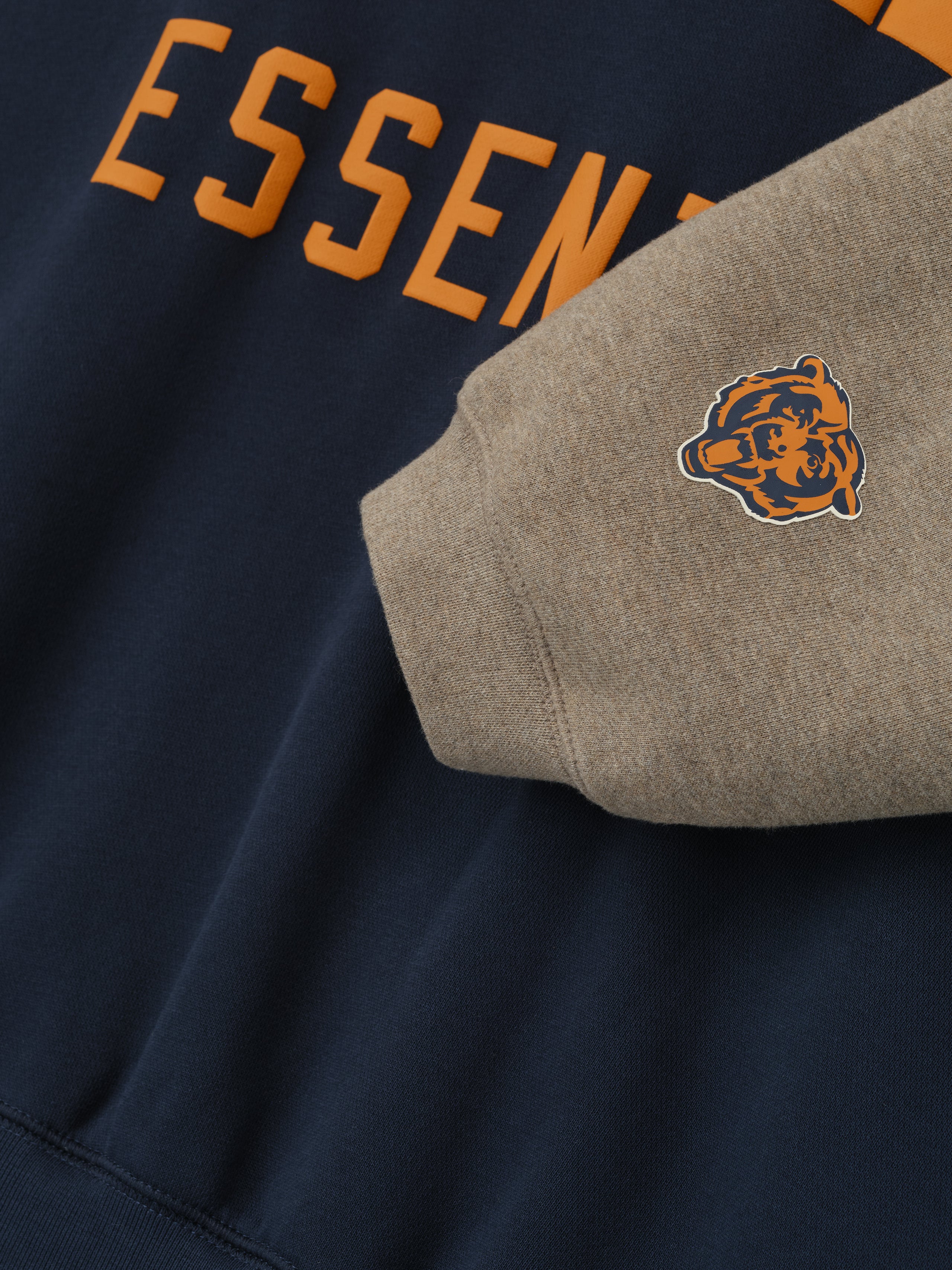 Close-up of Bears Sport Hoodie showing orange Essentials text on dark sapphire fabric and gray sleeve with small bear logo patch