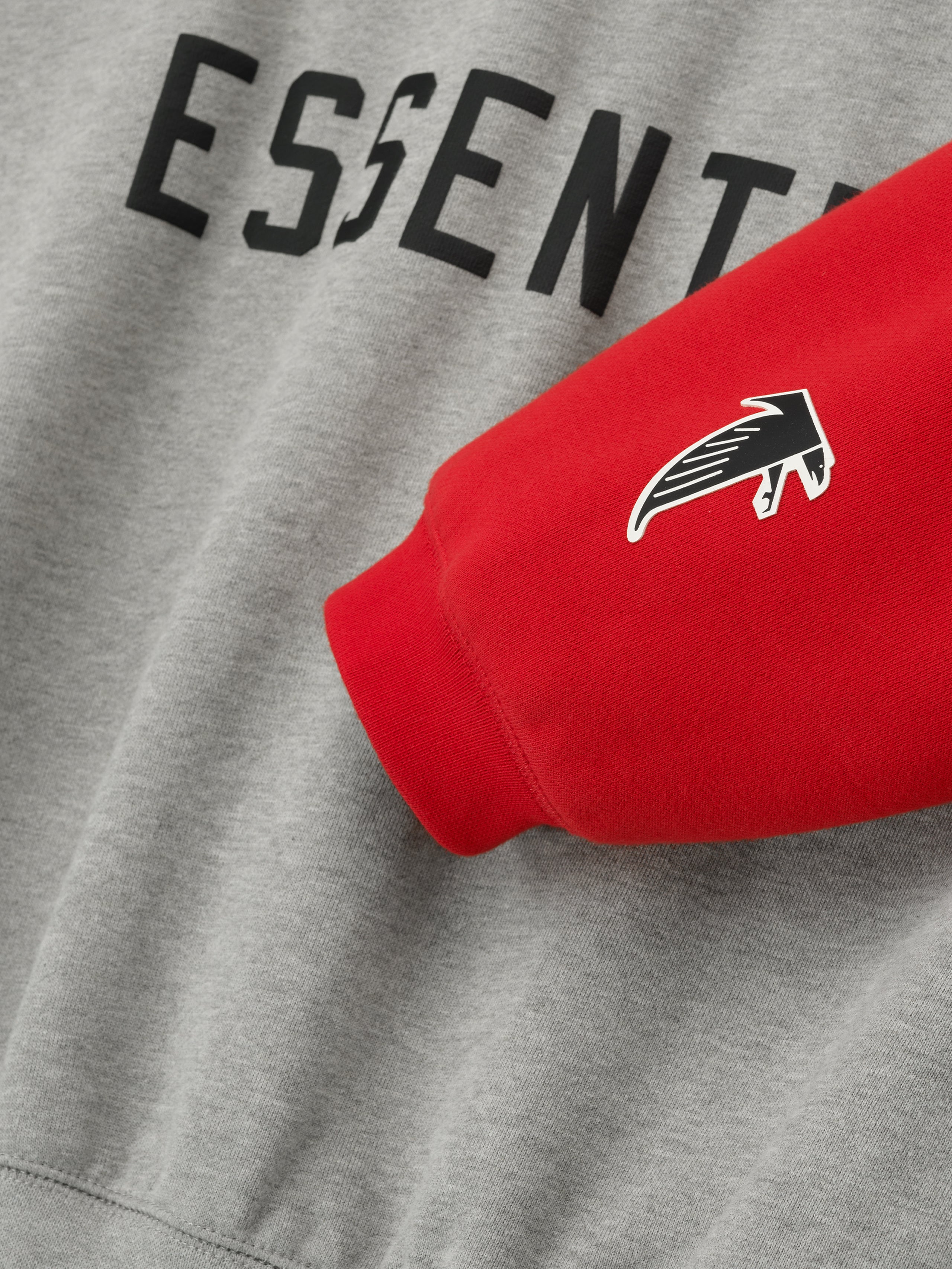 Close-up of Falcons Sport Hoodie showing red sleeve with team logo and black Essentials text on concrete heather fabric