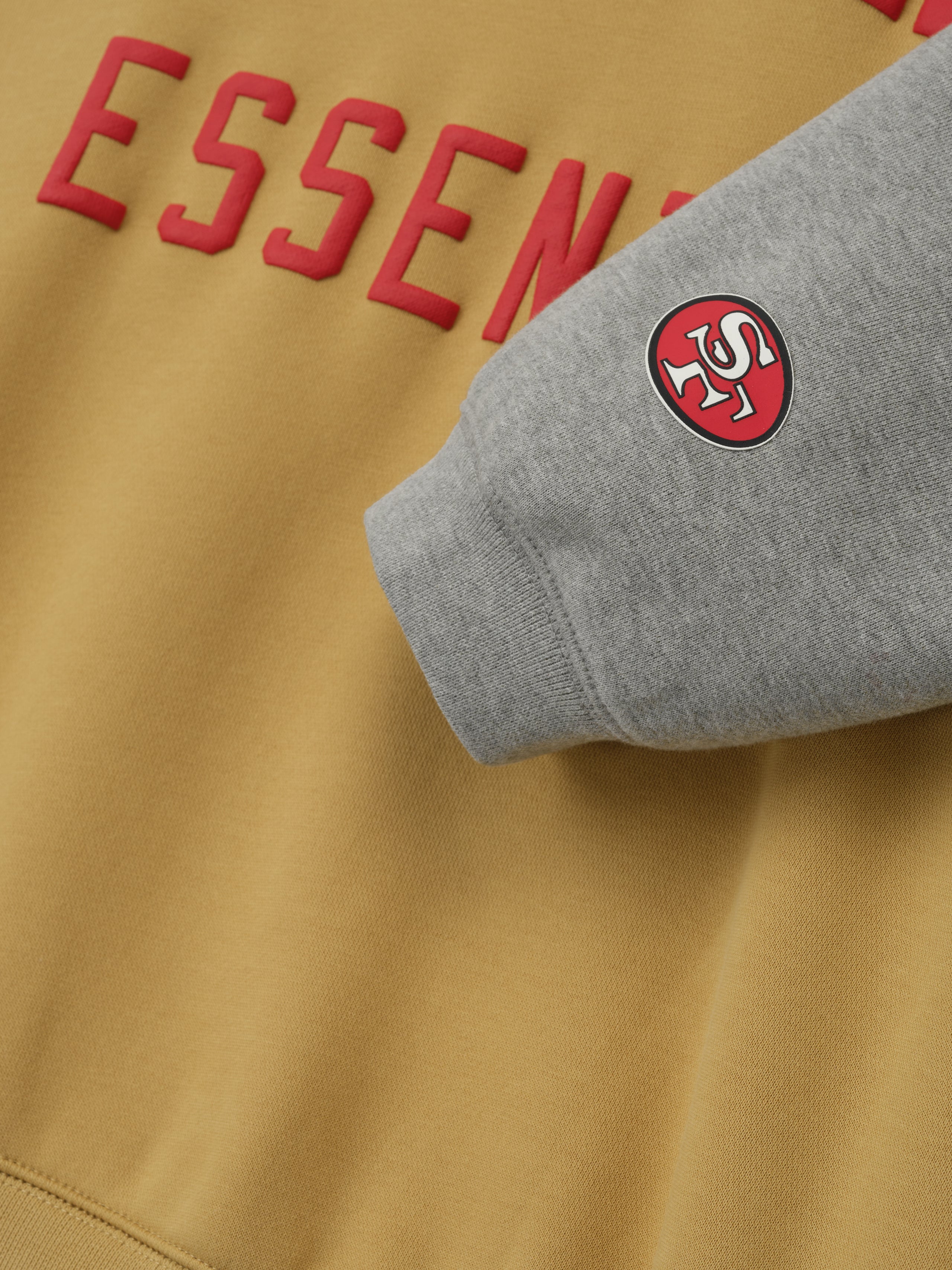 Close-up of Forty Niners Sport Hoodie showing red Essentials text on prairie sand fabric and gray sleeve with team logo patch