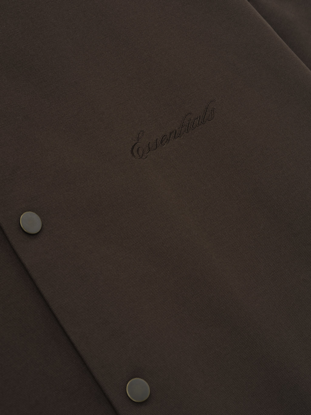 Close-up of dark brown fabric with tonal "Essentials" embroidered logo and two matte snap buttons on textured material