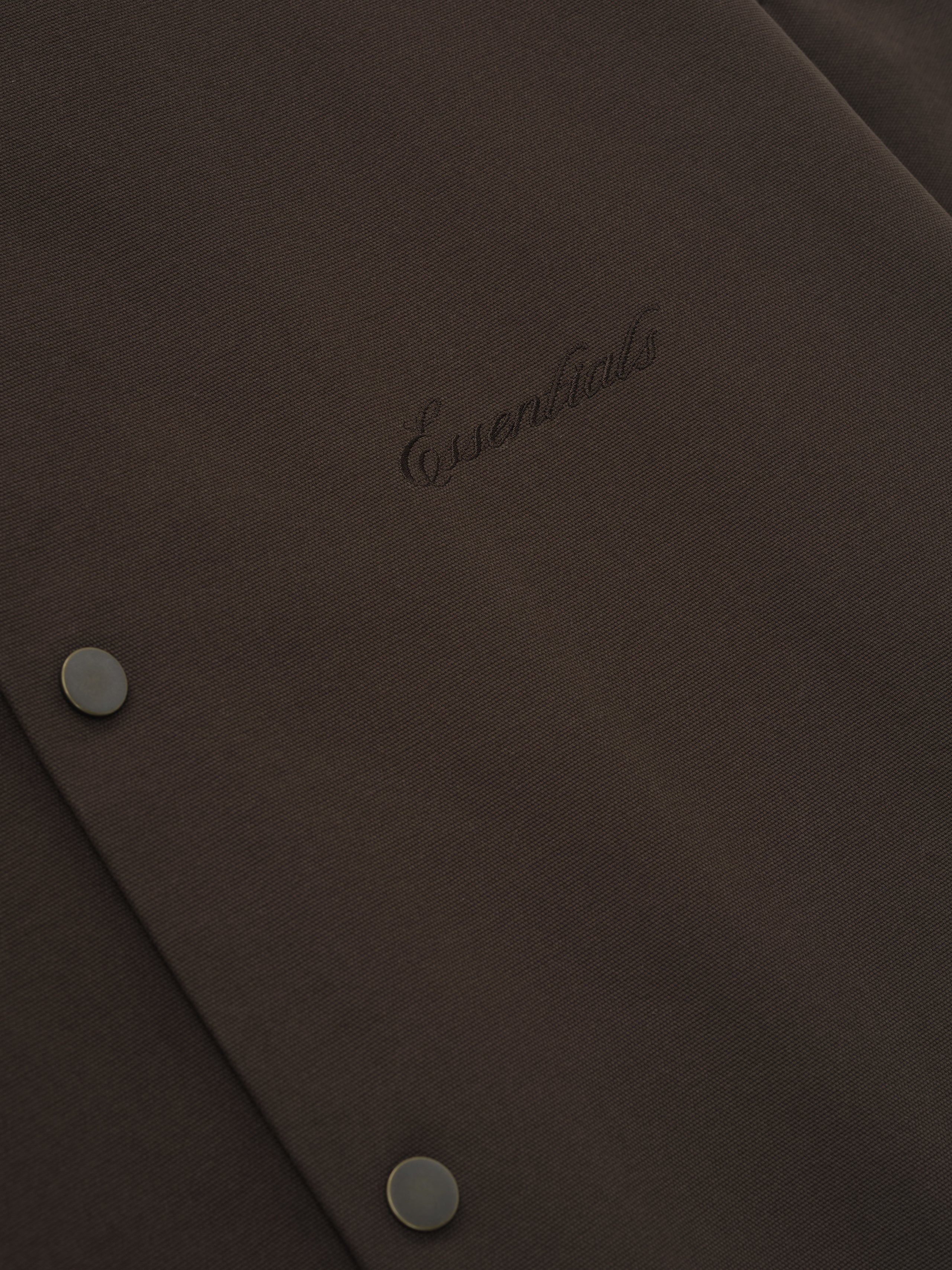 Close-up of dark brown fabric with tonal "Essentials" embroidered logo and two matte snap buttons on textured material