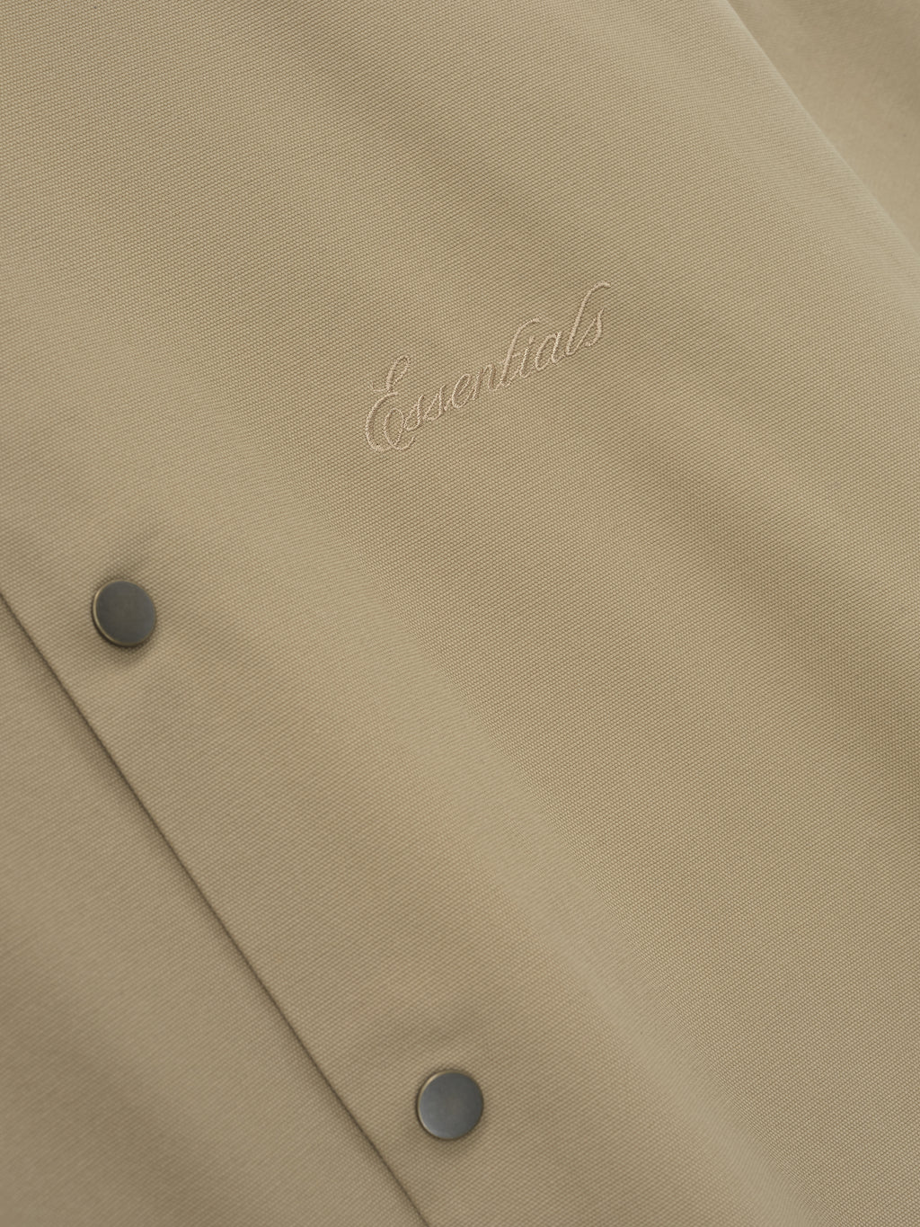 Close-up of beige fabric with tonal cursive "Essentials" embroidery and two matte snap buttons on textured material