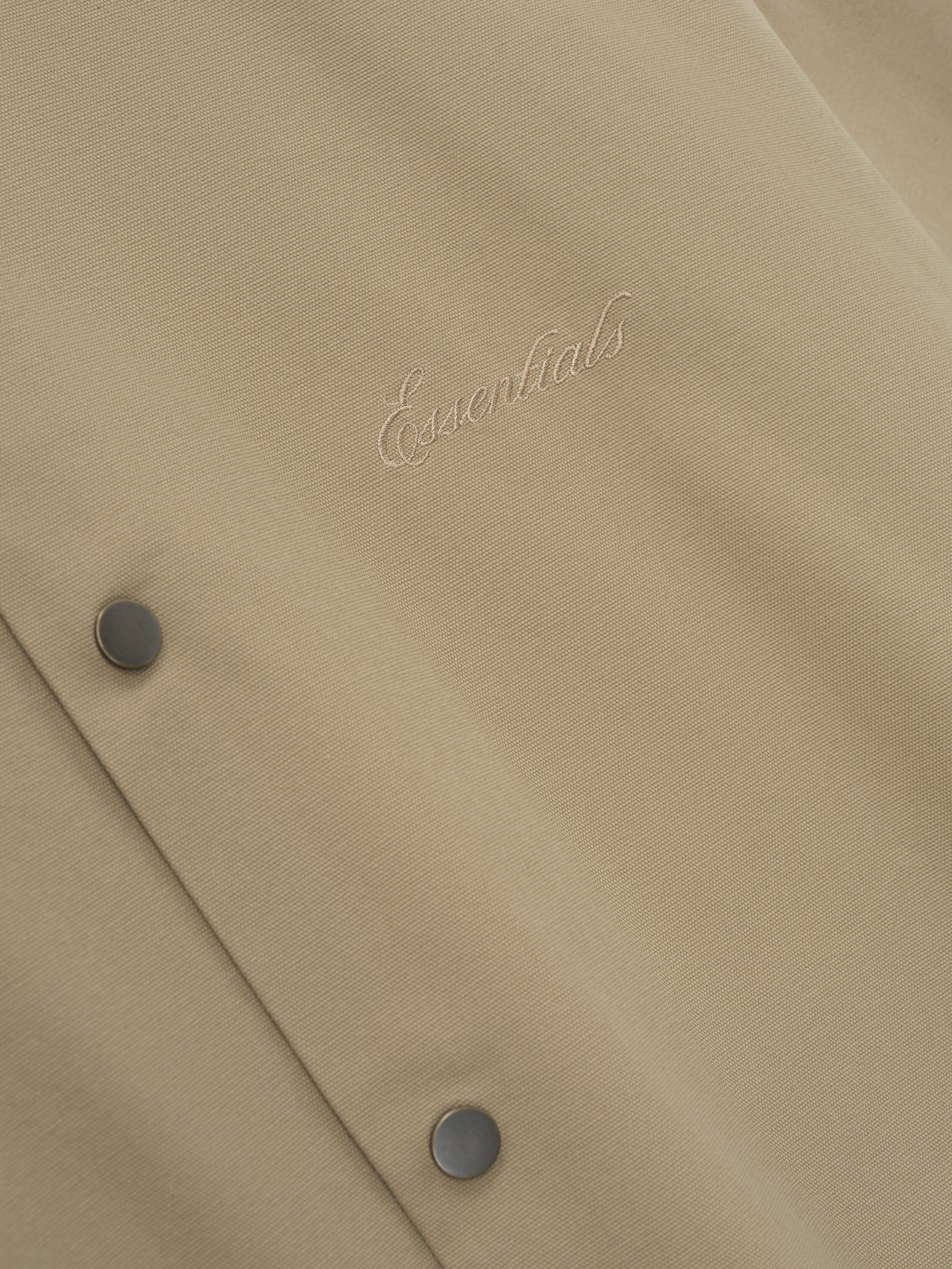 Close-up of beige fabric with tonal cursive "Essentials" embroidery and two matte snap buttons on textured material