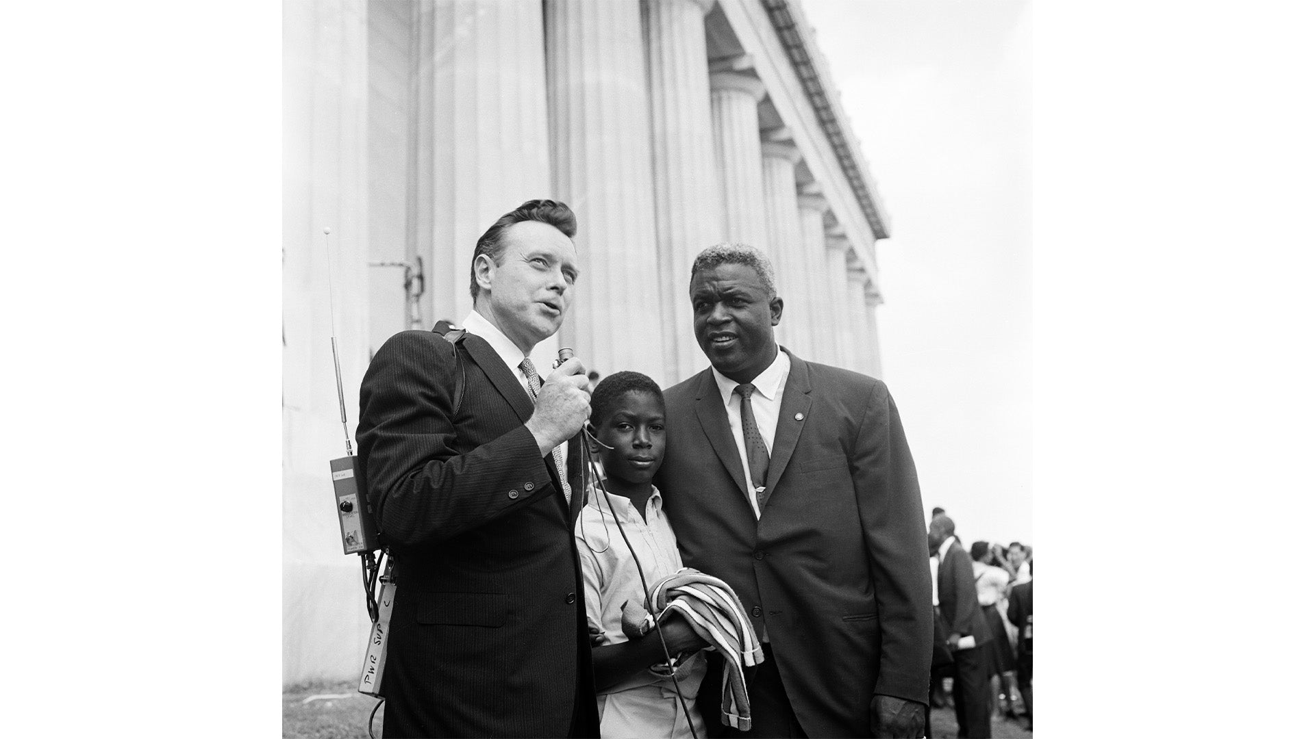 A black and white photograph of three men standing outside a building with tall columns, one man speaking into a microphone while the others listen attentively.