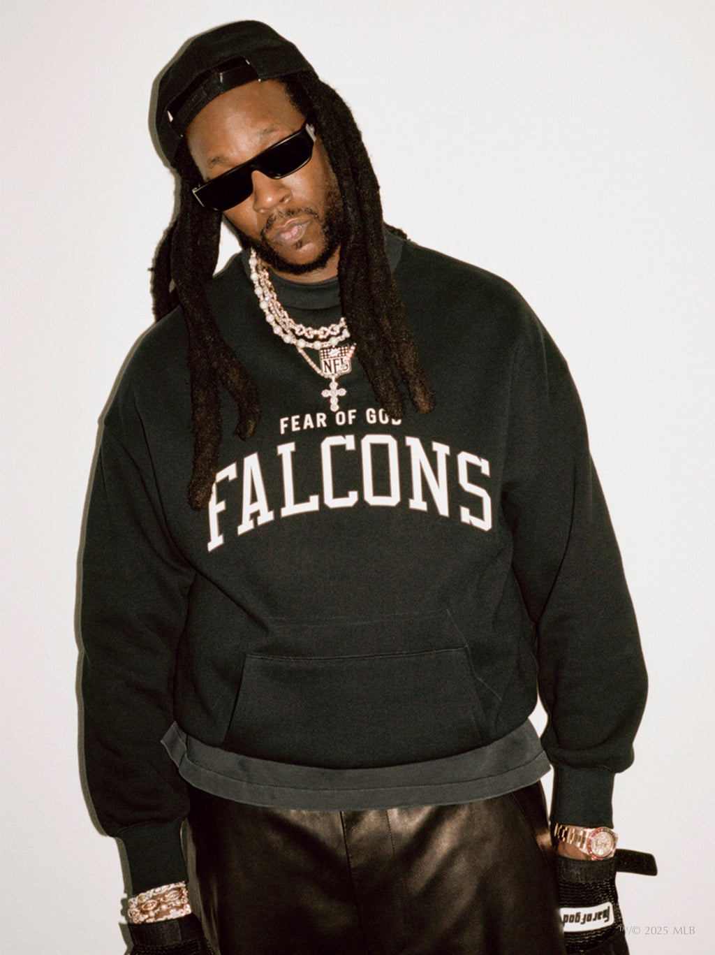 Man wearing black Essentials Falcons crewneck with white collegiate-style text and accessorized with chains and sunglasses