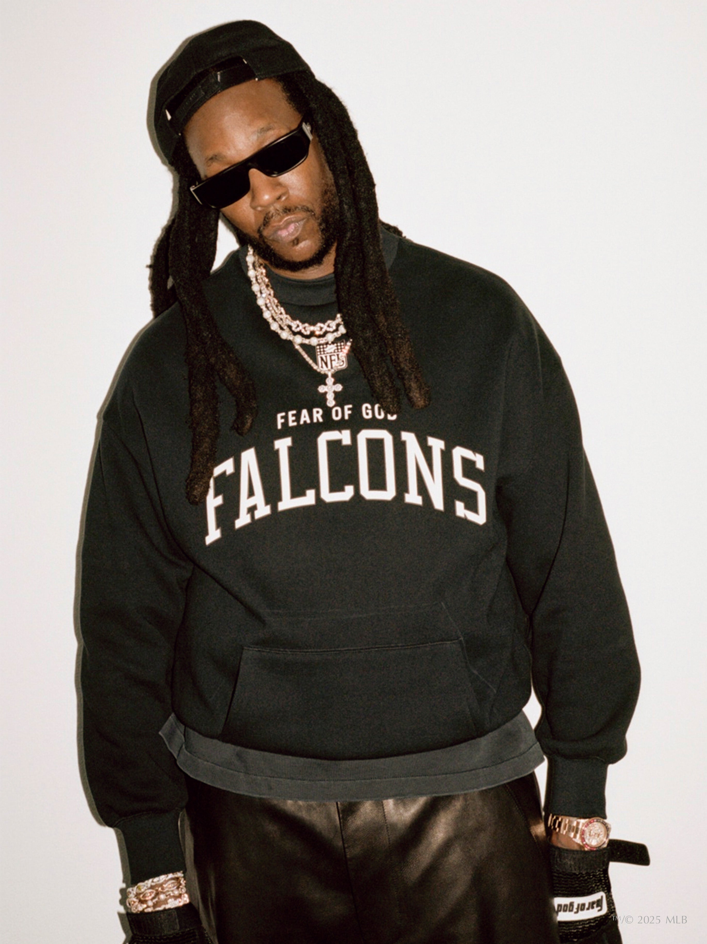 Man wearing black Essentials Falcons crewneck with white collegiate-style text and accessorized with chains and sunglasses