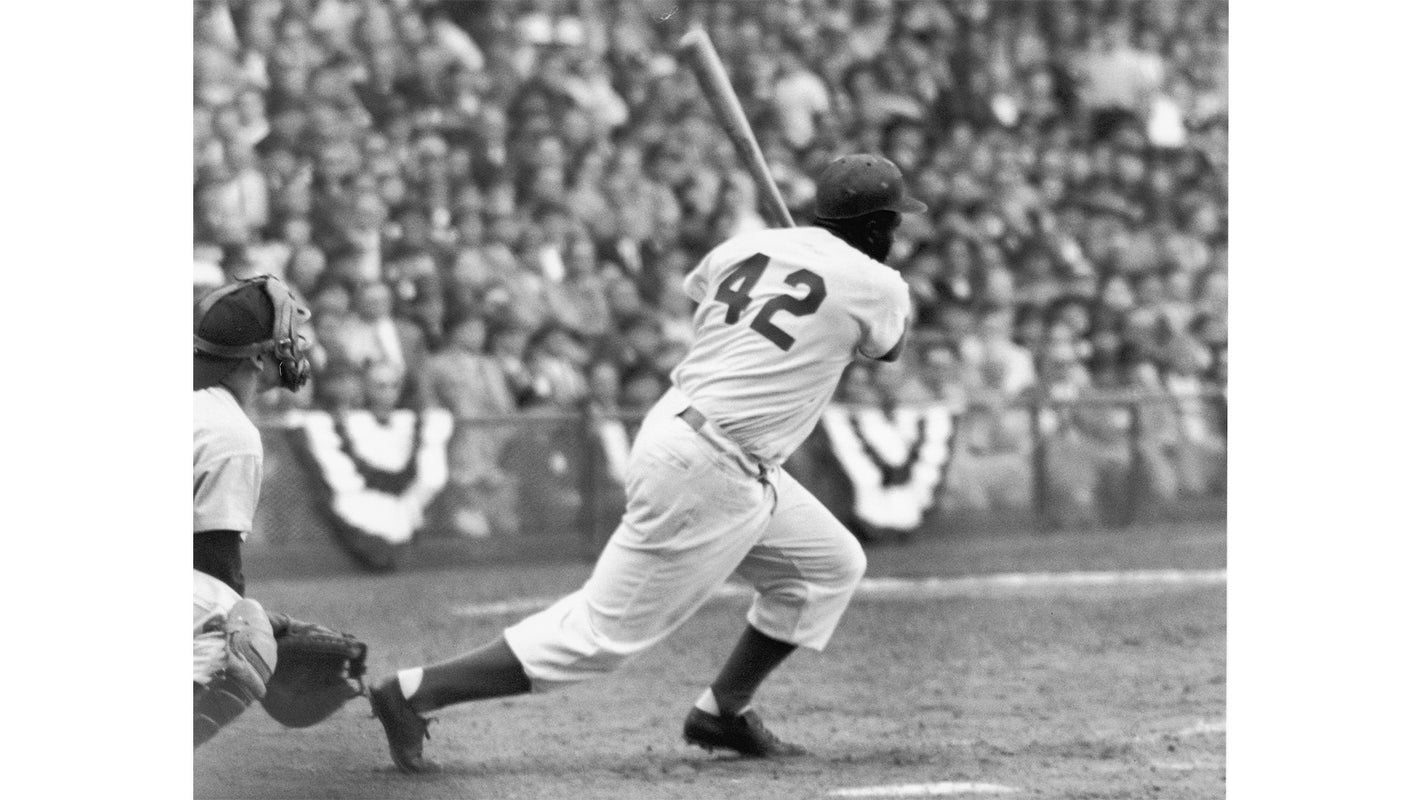 A black and white image capturing a baseball player wearing number 42 swinging a bat during a game, with a catcher positioned behind him and a crowd filling the stands in the background.