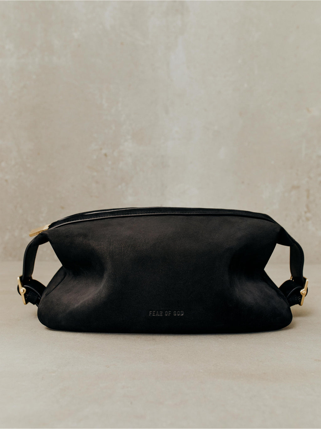 Black suede pouch bag with top zipper, side buckle straps, and subtle embossed branding on textured neutral background
