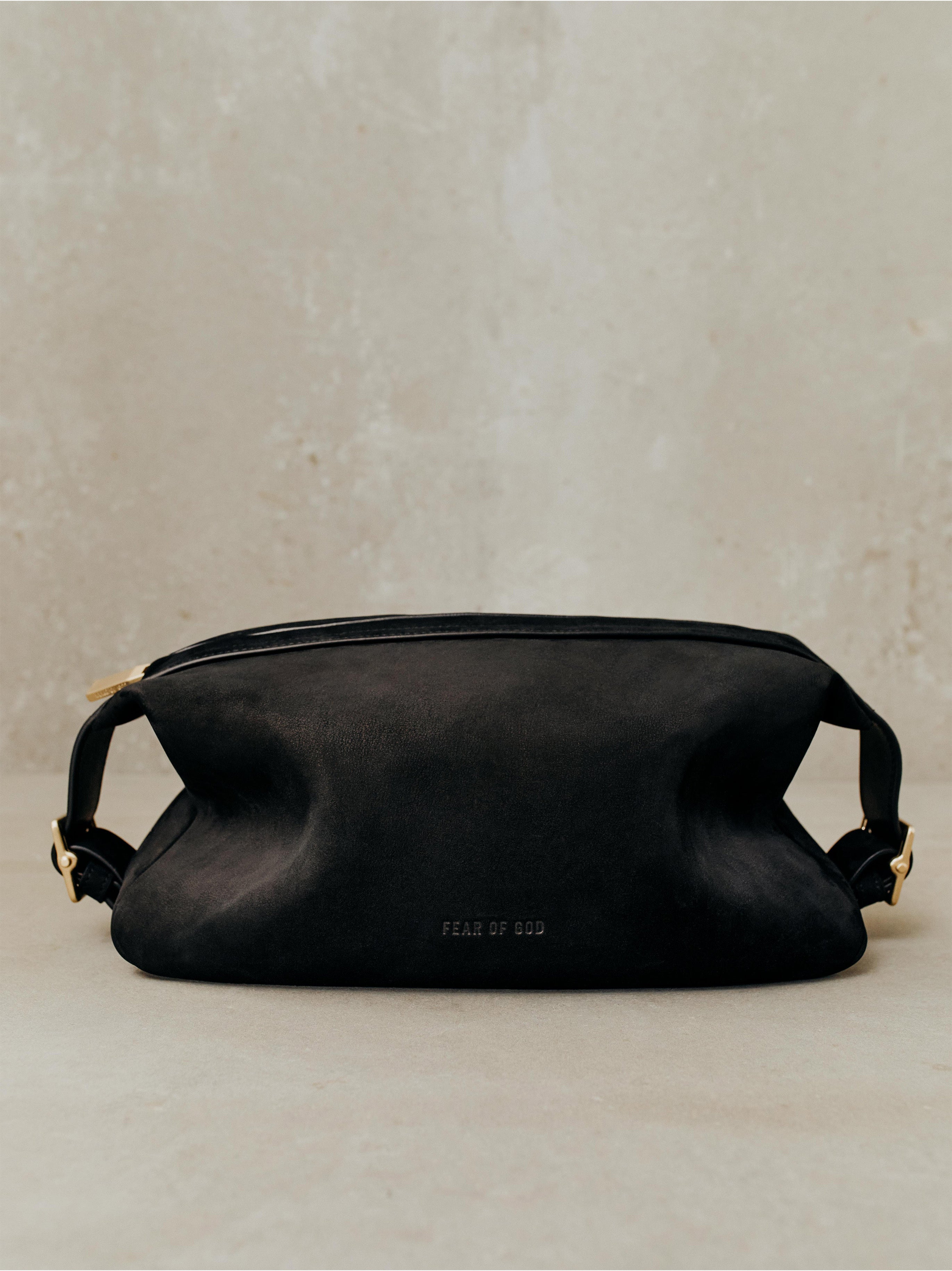 Black suede pouch bag with top zipper, side buckle straps, and subtle embossed branding on textured neutral background