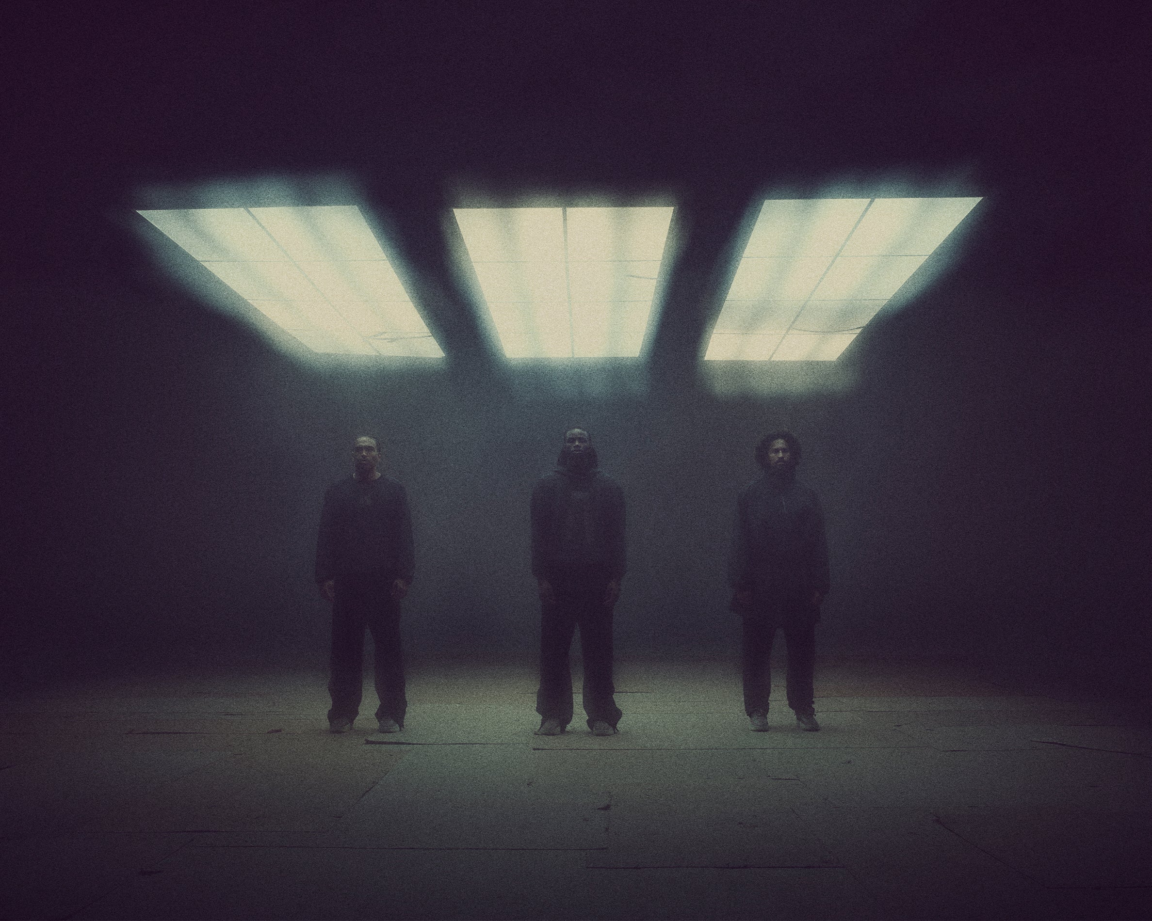 Three figures dressed in dark clothing stand spaced apart beneath three bright rectangular ceiling lights in a dim space