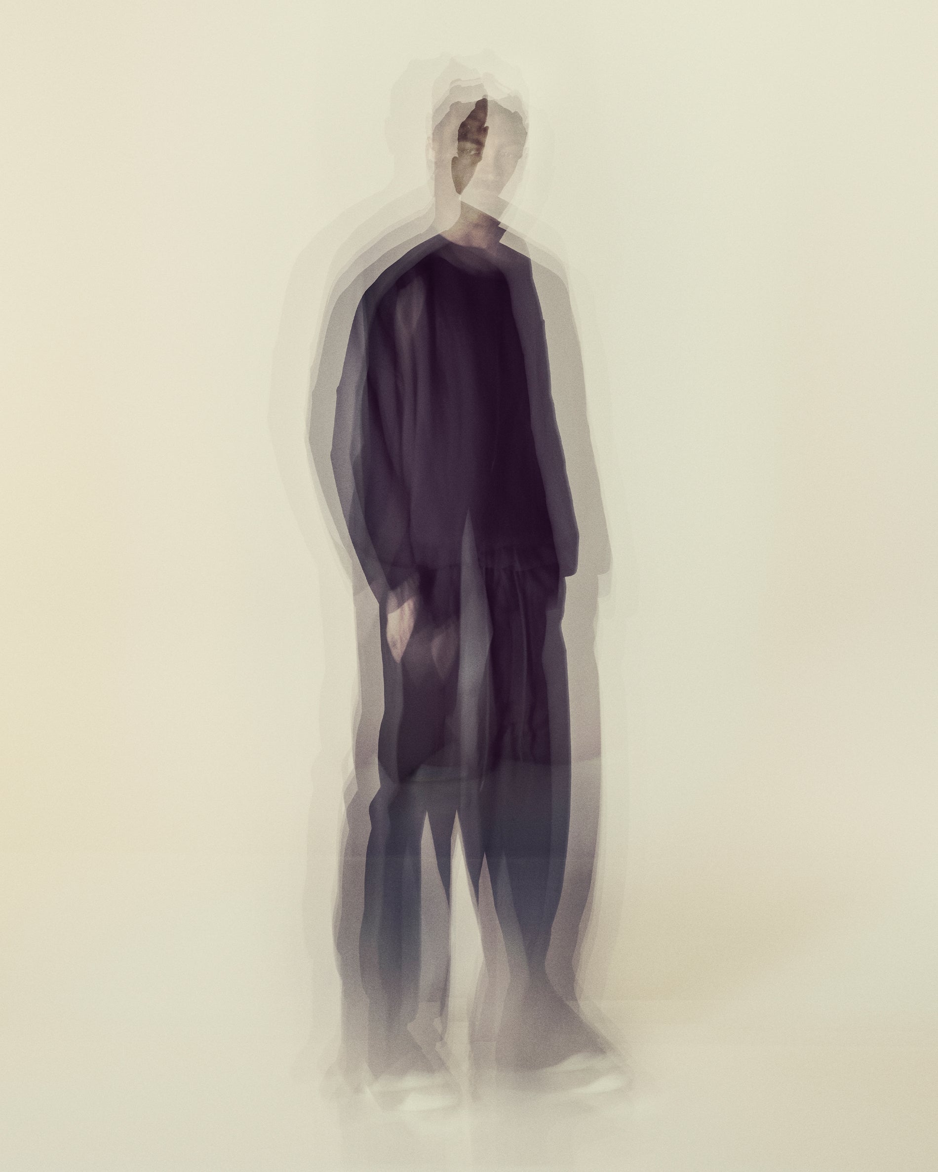 Blurred multiple exposure image of a person wearing dark loose clothing standing on a light background