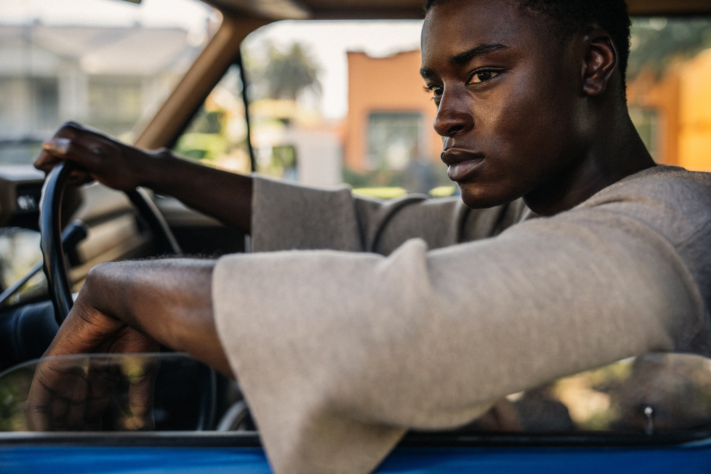 Young man with serious expression driving blue vintage car, resting arm on open window, city buildings in background