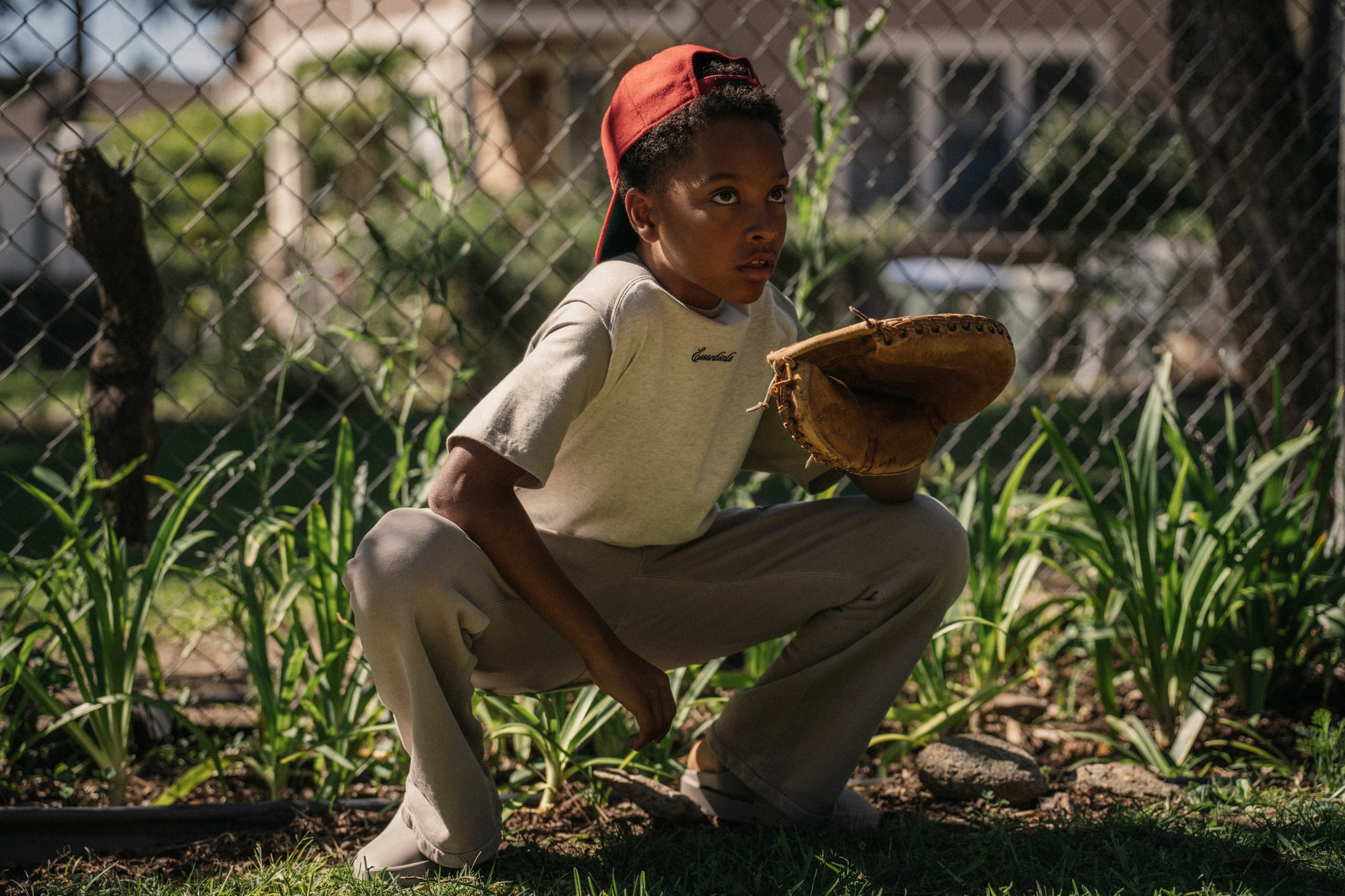 Boy wearing red cap backward squatting outdoors with baseball glove ready behind chain-link fence and grass