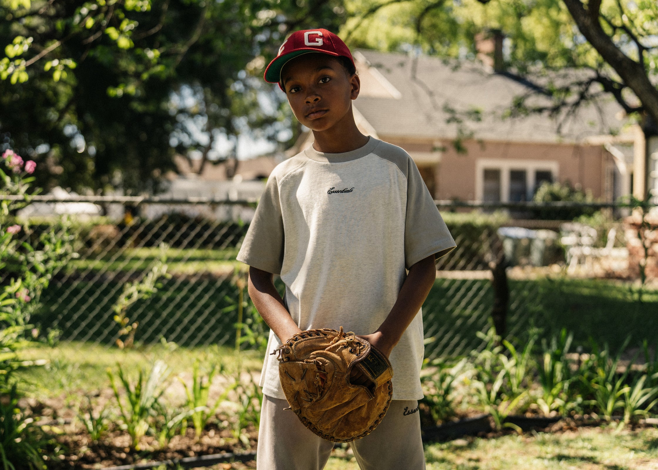 Young boy wearing red cap holding baseball glove standing in sunny backyard with greenery and house in background
